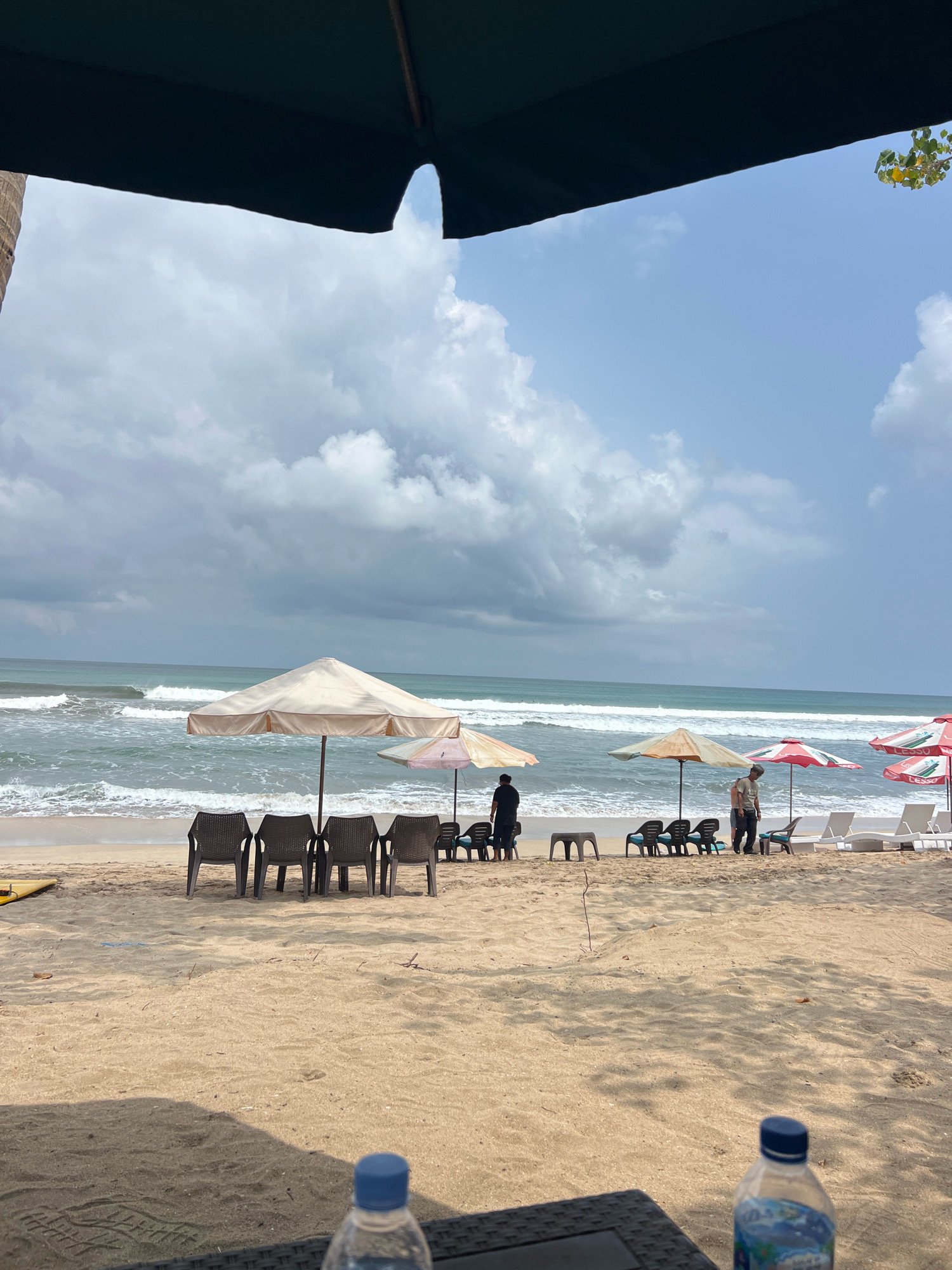 This is a photograph of a beach in Kuta, Indonesia, on a cloudy day. In the foreground, a dark umbrella partially obscures the view of the sky, which is filled with dramatic, gray clouds. The beach is sandy, and in the middle ground, several rows of plastic chairs are arranged facing the ocean. Some of these chairs are empty, while others are occupied by a few people. Large beach umbrellas, some a light tan color and others red and white, provide shade over seating areas. The ocean is visible with moderate waves breaking on the shore. In the background, the water stretches out to the horizon under the overcast sky. Two bottles of water are visible in the very foreground, placed on a dark, woven surface, likely a table.