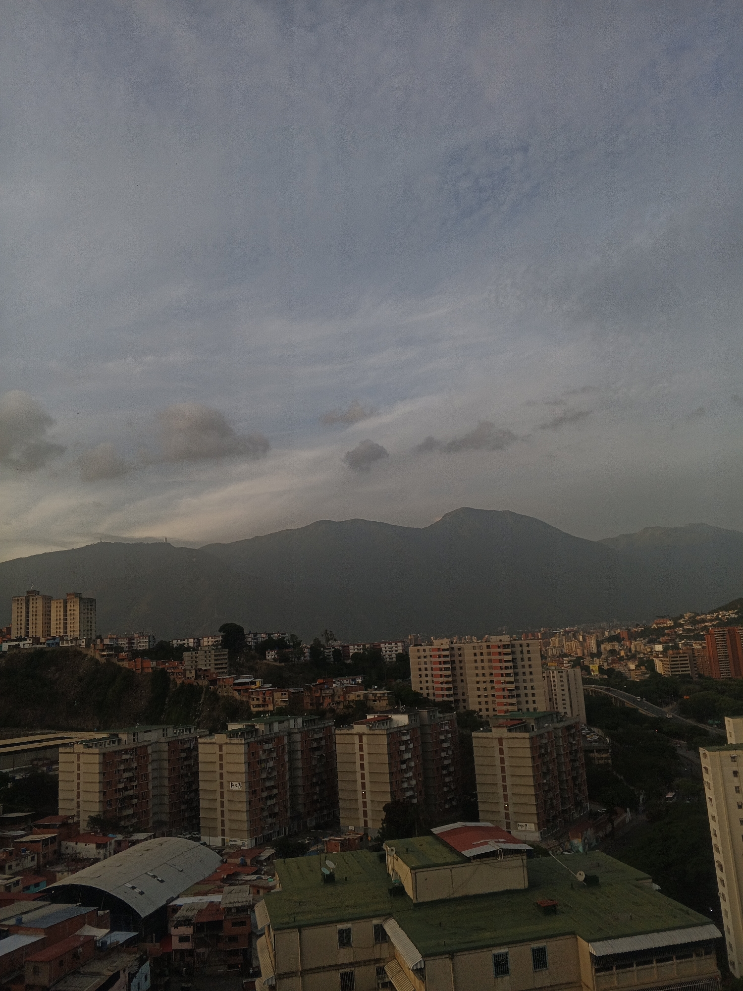 The image is a high-angle cityscape of Caracas, Venezuela, under an overcast sky. In the background, a mountain range dominates the horizon. The foreground and midground are filled with densely packed residential buildings, varying in height and architectural style. Some buildings have visible satellite dishes on their roofs. A highway is visible meandering through the urban landscape. No people are discernable in the image. The overall scene depicts a typical urban environment in Caracas.