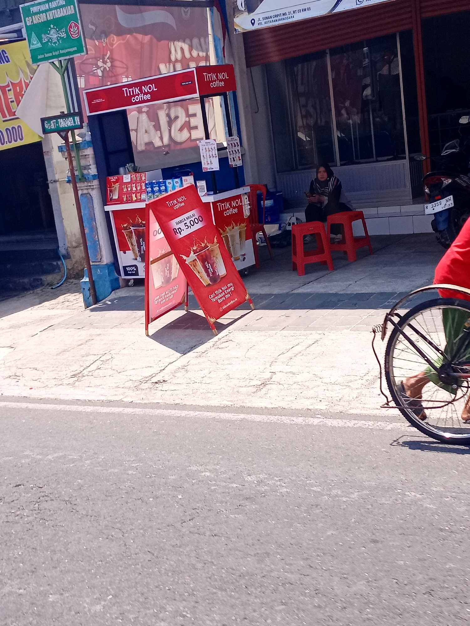 The image depicts a street scene in Banjarnegara, Indonesia, in front of a shop. A red and white A-frame sign for "TITIK NOL COFFEE" is prominently displayed on the sidewalk, advertising coffee with a price of Rp. 5,000. Behind the sign, a small kiosk or stall is visible, with shelves stocked with various drink products. A woman is seated on a red stool near the shop entrance, looking at her phone. Red stools are also placed outside the shop. A motorcycle with license plate R 3359 NM is parked to the right of the shop. In the foreground, the rear wheel and part of the frame of a bicycle are visible, suggesting someone is riding past the scene. The environment includes a paved sidewalk and a street. Overhead, a green sign with text that appears to be in Indonesian, including "PIMPINAN RANTING GP ANSOR KUTARANJARNEGARA", is mounted on a pole. Another yellow sign with text and a white banner is also visible. The shop's facade features large windows and a burgundy awning.