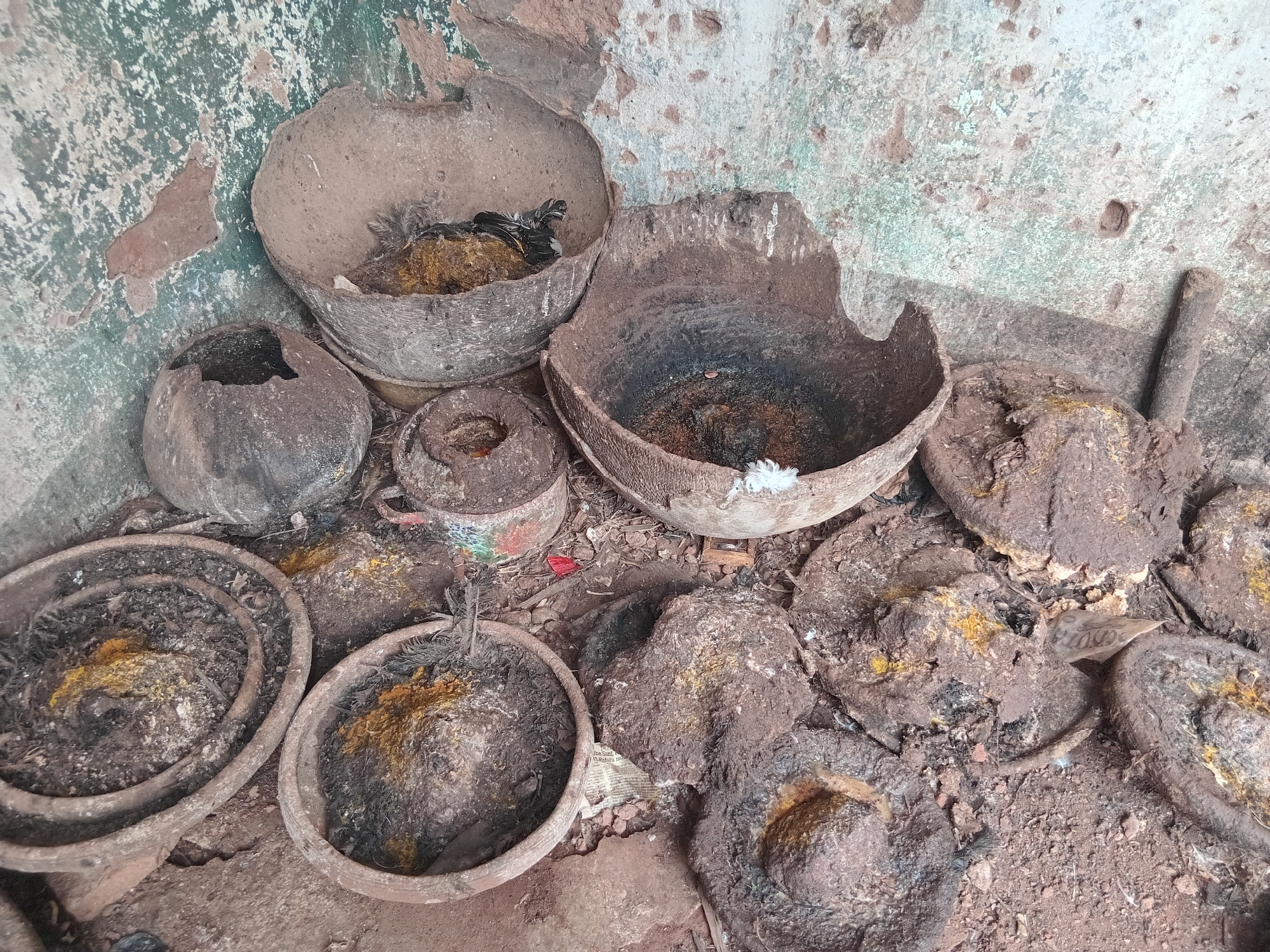 The image depicts an apparent ritualistic site in Shagamu, Nigeria, featuring numerous earthenware objects on a rough, earthy ground against a distressed wall with peeling greenish-white and reddish-brown paint.
The scene is dominated by various sizes of ceramic pots and broken bowls, many appearing aged, burnt, and encrusted with dark residues. Several of these containers hold burnt organic material and bright yellow or orange powdery substances, suggestive of traditional offerings or spices.
Notably, a broken pot in the upper left contains a small, dark, feathered bird carcass, also partially covered with the yellow substance, indicating a possible sacrifice. Another large, broken bowl in the center-right shows dark, burnt contents and a small white feather. Scattered around are several mud-like mounds, similarly adorned with the yellow powder. A rusted metal can or cup and a wooden stick are also visible among the items.
The presence of burnt offerings, the bird carcass, and the specific arrangement of objects strongly suggest a traditional religious or spiritual ritual has taken place or is ongoing. A small piece of newspaper visible on the ground in the lower right includes partially legible text such as "...Lagos na...", further reinforcing the Nigerian location context. The overall atmosphere conveys a sense of ancient practice and worn tradition.