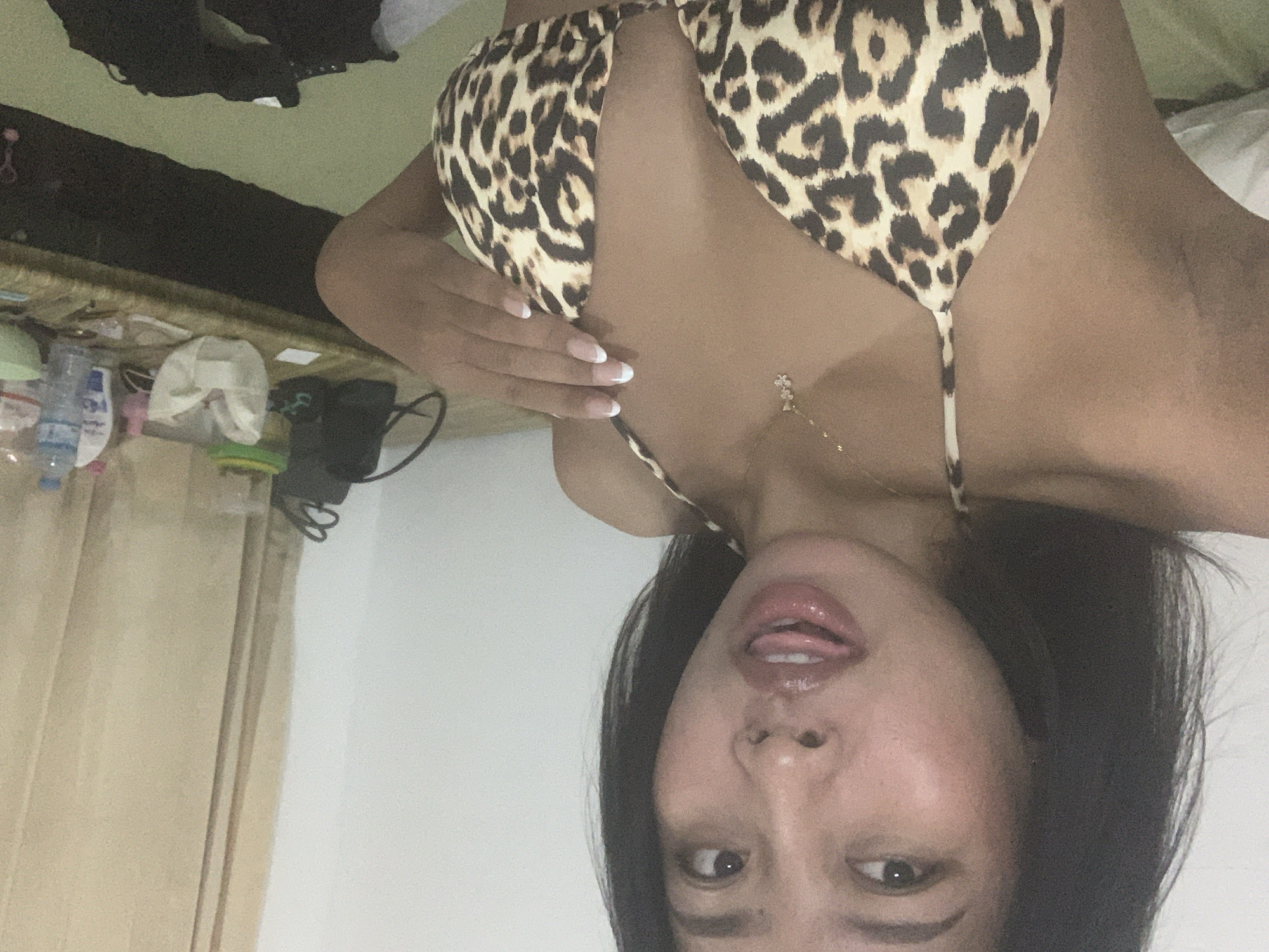 A woman with black hair and light brown skin wears a leopard-print bikini top, a gold necklace with flower charms, and has a French manicure. She is sticking her tongue out and looking directly at the camera. The scene takes place indoors, with a cluttered desk visible in the background. A white curtain is visible on the left. The location cannot be determined, as there are no geographical landmarks or signs indicating that the image was taken in Denpasar, Indonesia.