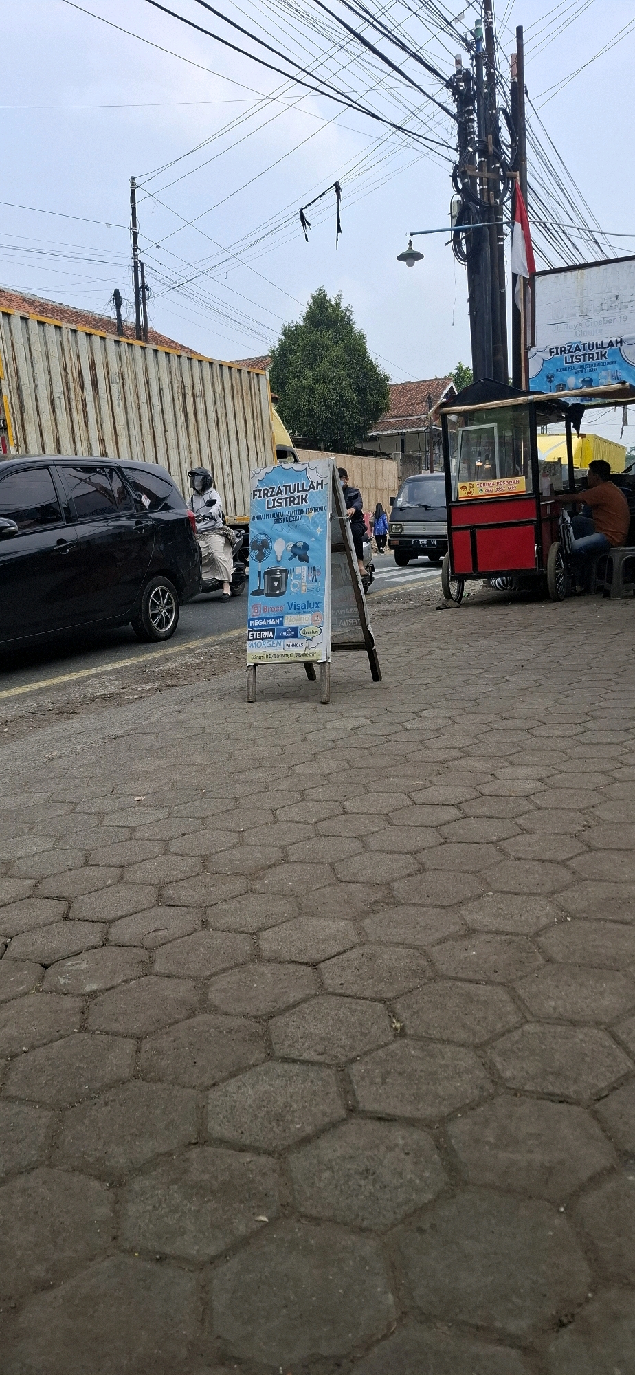 The image depicts a street scene in Sirnagalih, Indonesia. In the foreground, a cobblestone pavement is visible. A black minivan is parked on the left side of the road, with a person on a motorcycle behind it. To the right, a red food cart with a glass display is parked, and a person is seated behind it. An A-frame sign with blue and white text advertising "FIRZATULLAH LISTRIK" (electricity) is positioned in the center of the frame. The sign displays images of electrical appliances such as a fan, a light bulb, and a blender, along with brand names like "Brace," "Visalux," "Megaman," "Piolino," "Eterna," and "Morgen." In the background, a large metal container truck is parked, and a lush green tree stands between buildings with red tile roofs. Overhead, a complex network of electrical wires crisscrosses the sky, supported by utility poles. A street lamp is visible hanging from one of the poles. A red and white Indonesian flag is attached to one of the poles. The sky is overcast.