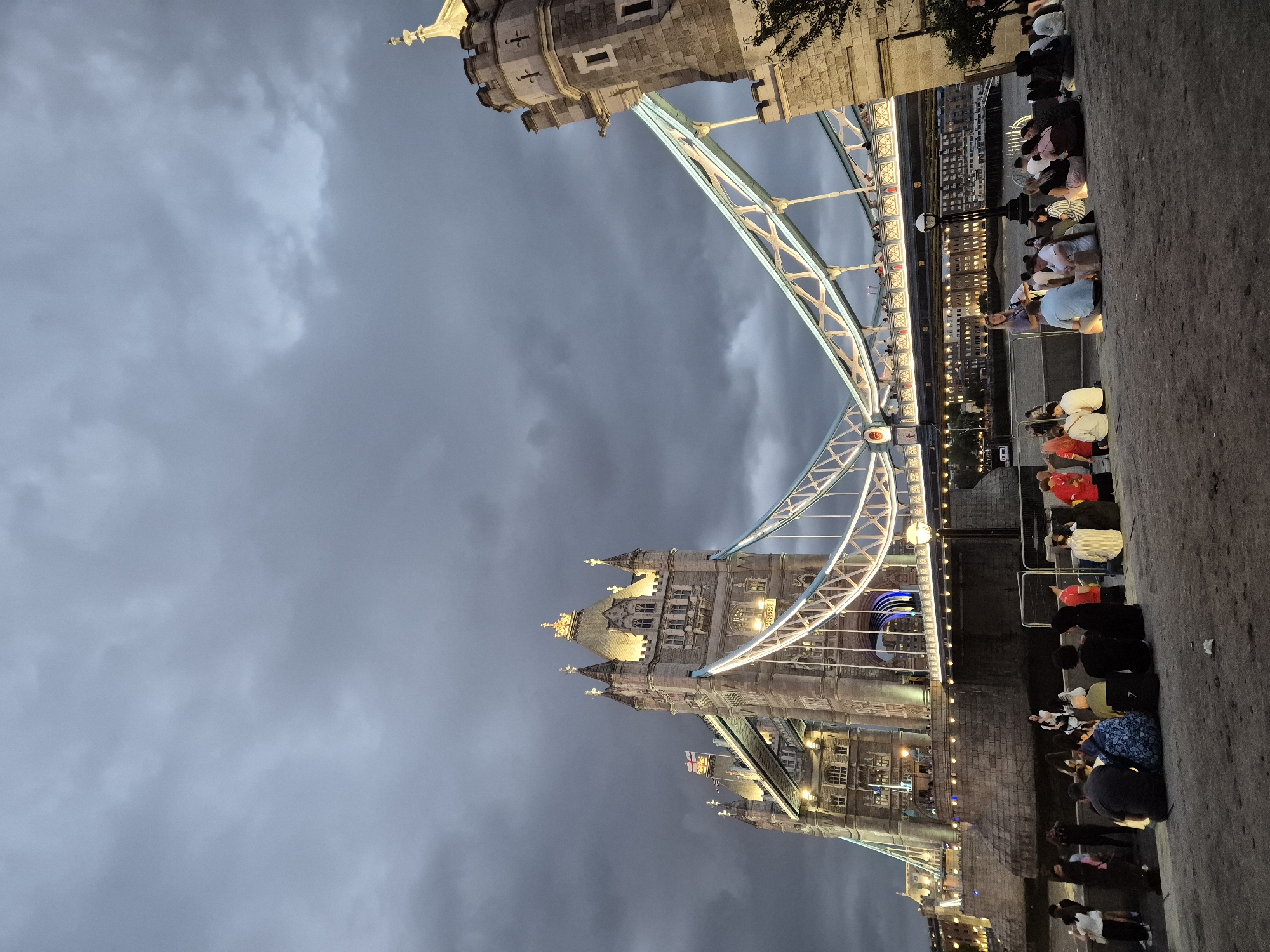 The image depicts Tower Bridge in London, United Kingdom, under a cloudy sky during dusk. A group of people are seated on the ground in the foreground, facing the bridge. They appear to be enjoying the view or gathered for an event. The bridge itself is illuminated, highlighting its architectural features. The surrounding area seems to be a public space along the Thames River. The overall mood is calm and somewhat atmospheric due to the lighting and the approaching evening.