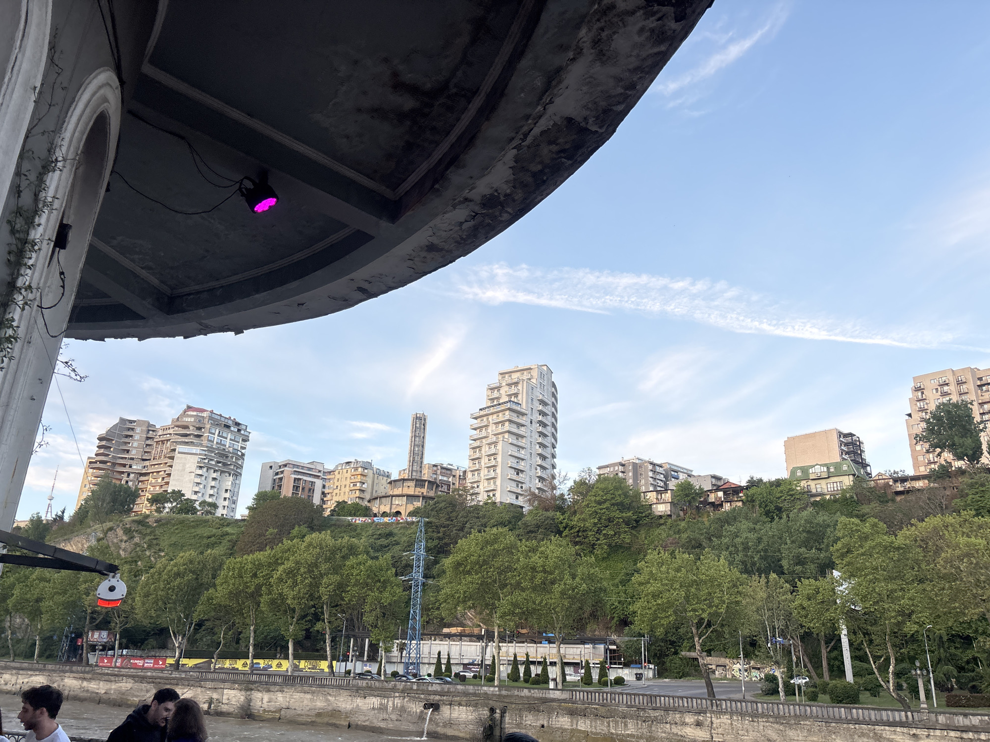 The image is a low-angle shot of a cityscape in Tbilisi, Georgia, taken from under a concrete overhang. In the foreground, a group of people are gathered, though their actions are not clearly discernible. The middle ground features a retaining wall and a tree-lined embankment. Above the embankment, a steep, green hill is dotted with a variety of buildings, including modern high-rises and older residential structures. A blue electrical tower is visible amidst the trees. In the background, the sky is light blue with wispy clouds. The concrete overhang in the foreground is a prominent architectural feature, with a pink light fixture attached to its underside.