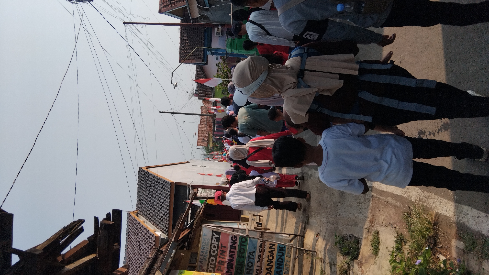 The image captures a street scene in Posong, Indonesia, likely during a daytime event or parade. A group of people are walking down a paved street lined with buildings on both sides. The buildings appear to be houses and small businesses, some with tiled roofs and white walls. Indonesian flags, red and white, are prominently displayed along the street, suggesting a national celebration or commemoration.

The crowd is diverse, with men, women, and children of various ages. Many are wearing casual clothing, while some women are wearing hijabs. The people are walking in the same direction, facing away from the camera, creating a sense of movement and procession. In the foreground, a young boy in a white t-shirt and black pants is walking, with other individuals behind him. To the left, a wooden stand with colorful signage is visible, displaying text that appears to be related to printing services. The sky above is clear and bright. Electrical wires are strung across the street, typical of urban or semi-urban environments in Indonesia. The overall atmosphere suggests a community gathering.