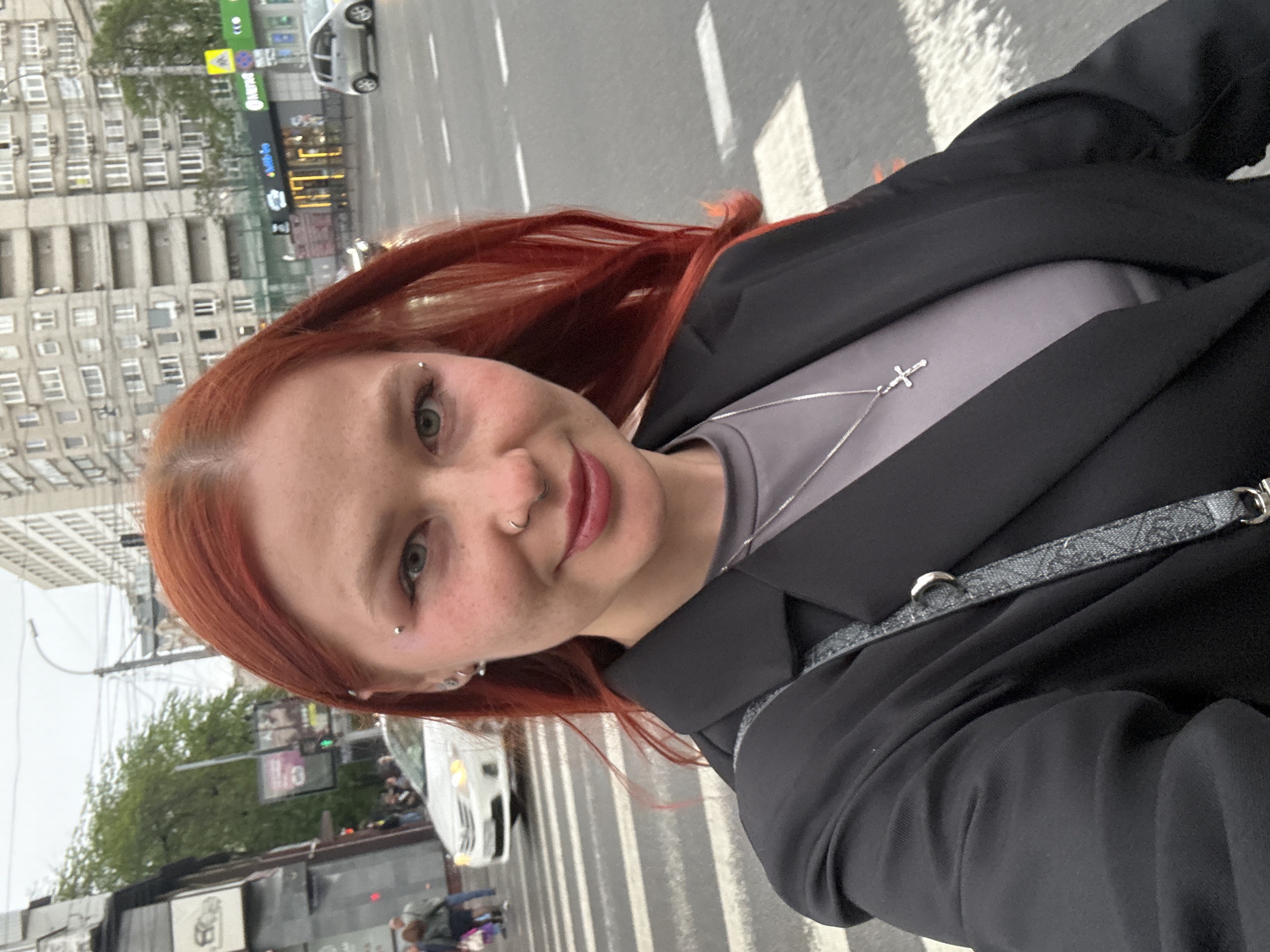 A woman with red hair and multiple piercings is taking a selfie on a crosswalk. She has piercings in her nose and eyebrow, and her hair is a vibrant red. She is wearing a black blazer over a gray shirt and a necklace with a cross pendant. A patterned strap, likely from a bag, is visible across her body.
In the background, there is a street with a white car crossing, a tall apartment building with multiple windows, and trees. There is a sign for a subway station called "Rostov-Glavniy" visible in Cyrillic. Given the subway station name, the location is likely Rostov-on-Don, Russia. Pedestrians can be seen walking on the sidewalk near the crosswalk.