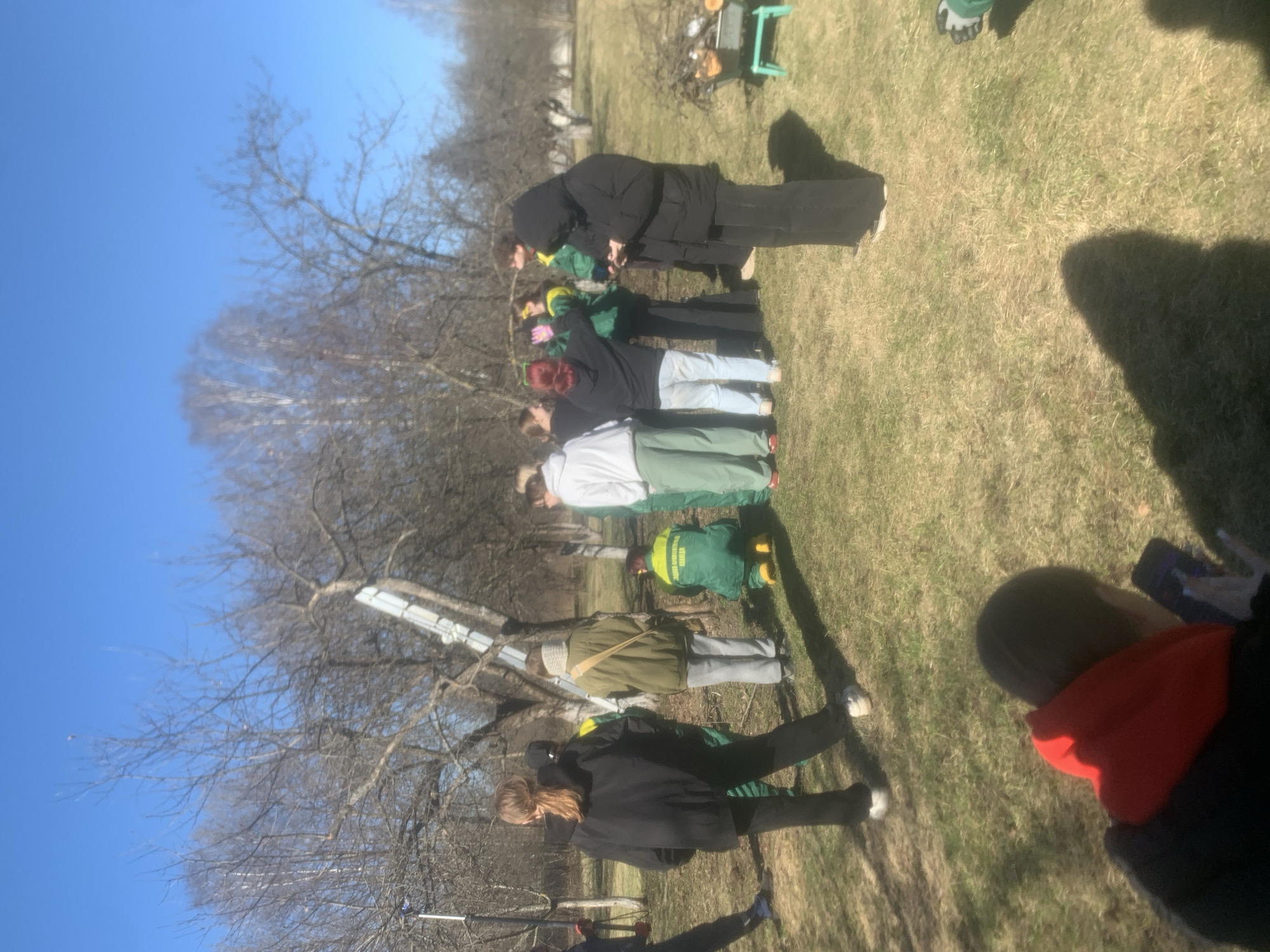 A group of people, including what appears to be children, are gathered in a grassy area on a sunny day. The location is likely outdoors, possibly a park or field. Bare trees indicate it is likely late fall, winter or early spring. A ladder leans against a tree. The group of people is standing in line. The city is mentioned as Saint Petersburg, Russia.