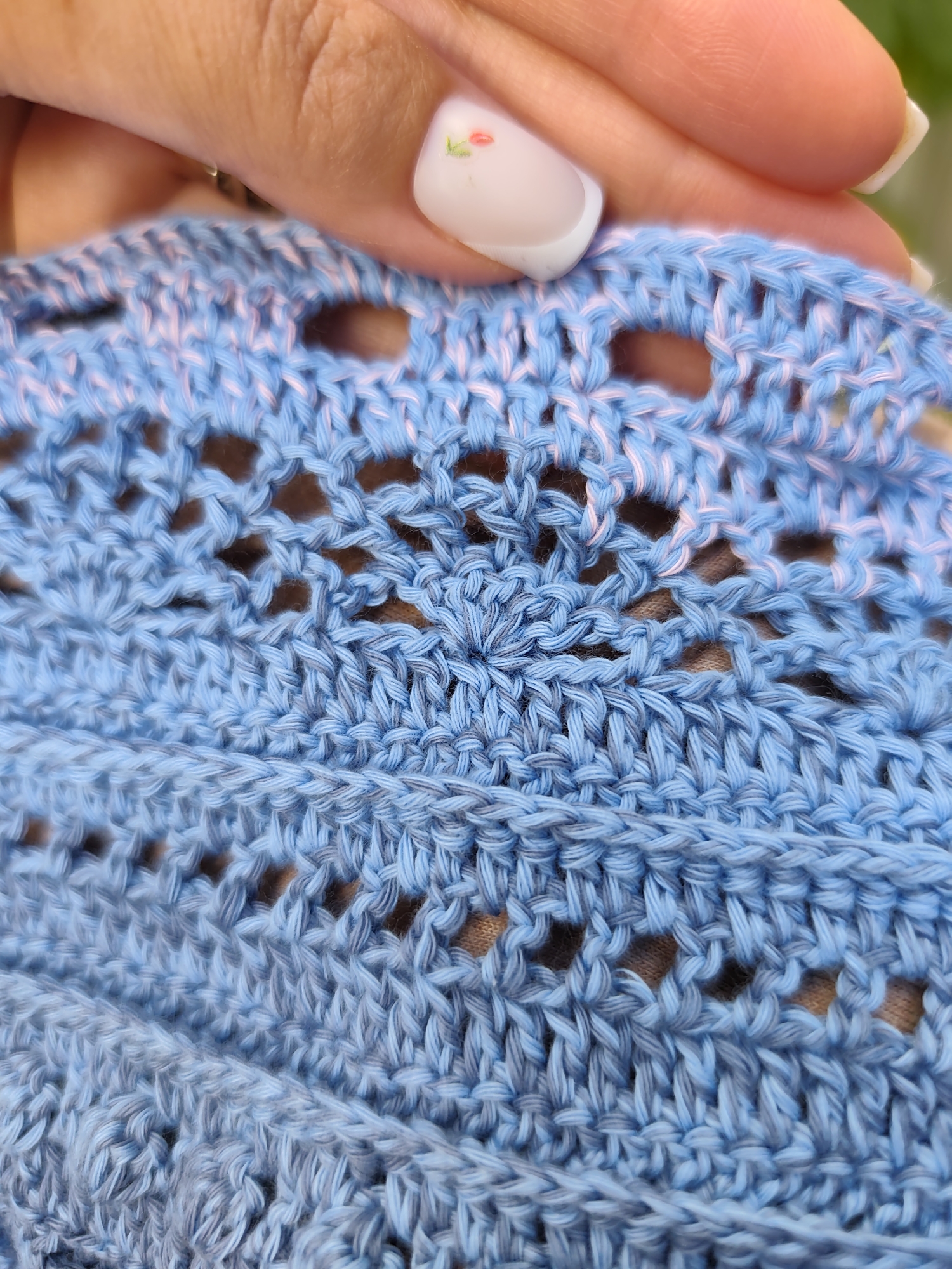 This is a close-up, medium shot photograph of a person's hand holding a piece of light blue crochet fabric. The hand is positioned in the upper right quadrant of the frame, with the fingers delicately touching the fabric. The fingernails are painted white with a subtle, light pink floral design on the thumbnail and index finger. The crochet fabric itself features a delicate openwork pattern with some pink and light gray threads interspersed within the predominantly blue yarn. The lighting suggests an outdoor setting, as indicated by the soft, natural light and the blurred green background, which could be foliage. The image's focus is on the texture and detail of the crocheted material. There is no visible indication of the location being in the city of Durlești, Moldova.