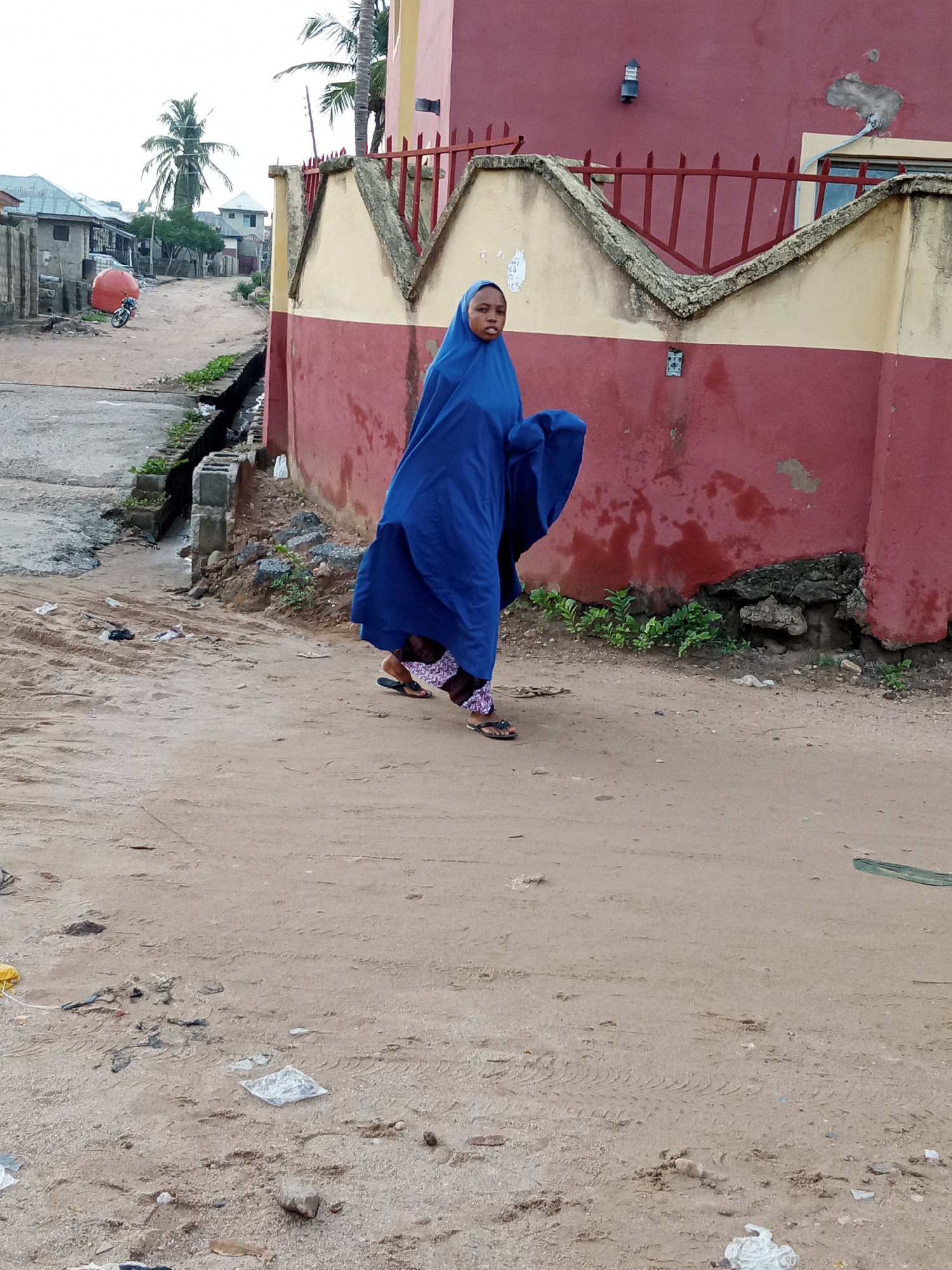 A person in a blue garment walks along a dirt road in Zango-ndaji, Nigeria. The road is sandy and contains debris. To the left is a drainage ditch and to the right is a building with a red and yellow wall topped with a red metal fence. In the distance are buildings and palm trees.