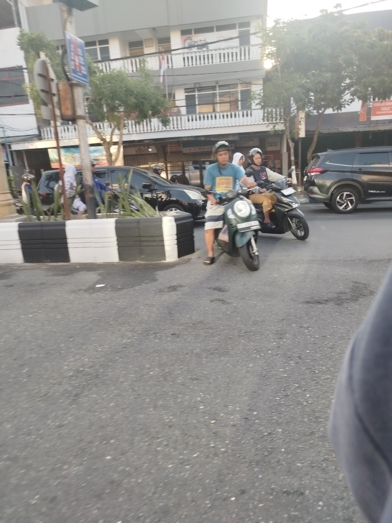 In Balikpapan, Indonesia, a street scene unfolds with two men on motorcycles. The man in the foreground, wearing a blue t-shirt and shorts, is on a dark green scooter. Behind him, another man in a white and red jacket rides a black motorcycle. Both men are wearing helmets. A black SUV is visible to the right, and other vehicles and buildings are present in the background. A black and white striped barrier is in the middle ground, separating the road from a planter with greenery. The lighting suggests it is daytime, possibly late afternoon due to the warm tones in the sky.
