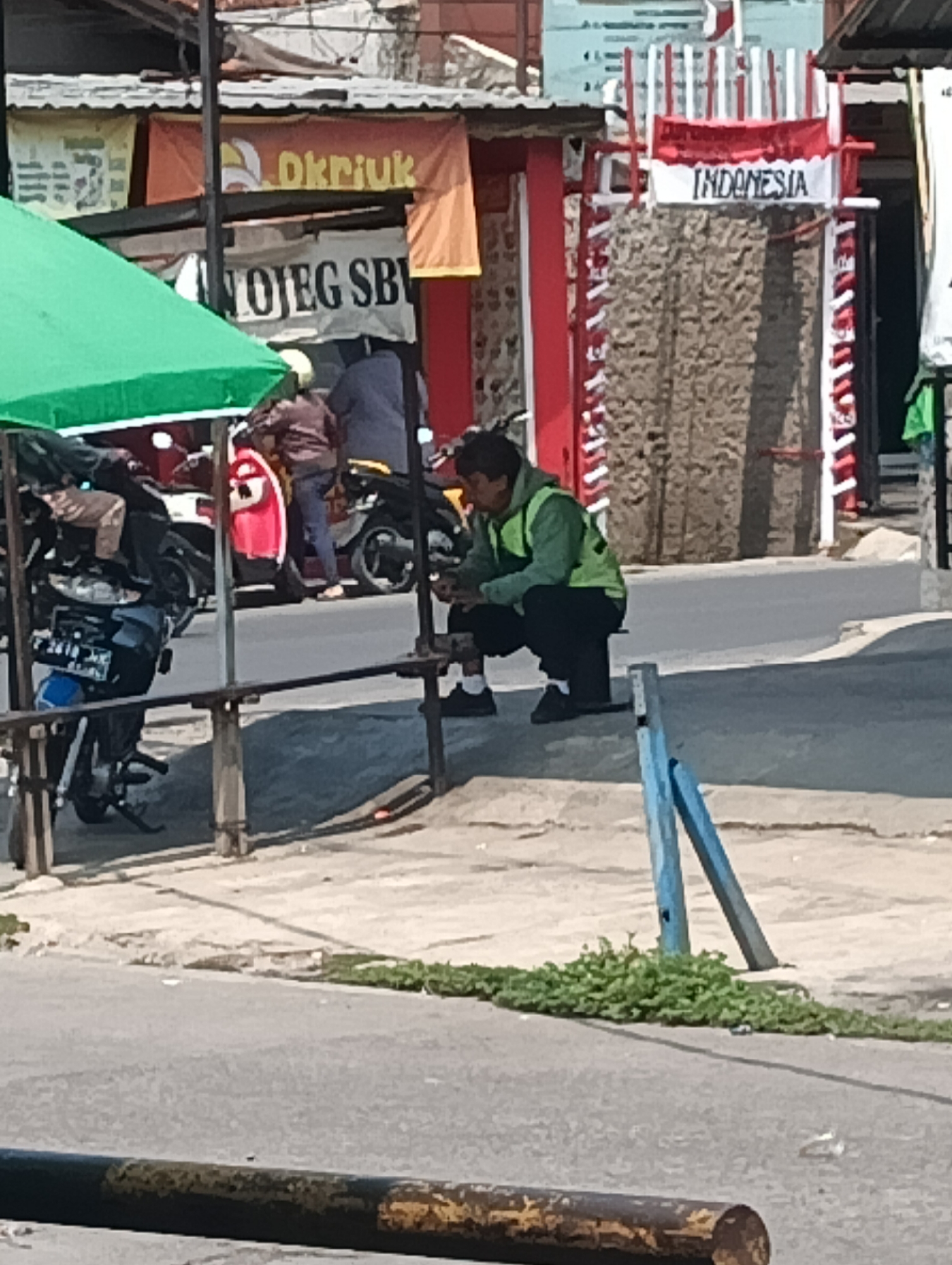 In Gandasari, Indonesia, a person wearing a bright green vest and dark pants crouches on a sidewalk near a street. The person appears to be looking down at something in their hands. Behind them, several motorcycles are parked, and a red and white banner with the word "INDONESIA" is visible on a structure. Shops and buildings line the street, with awnings providing shade. The scene is illuminated by daylight, suggesting it is daytime.