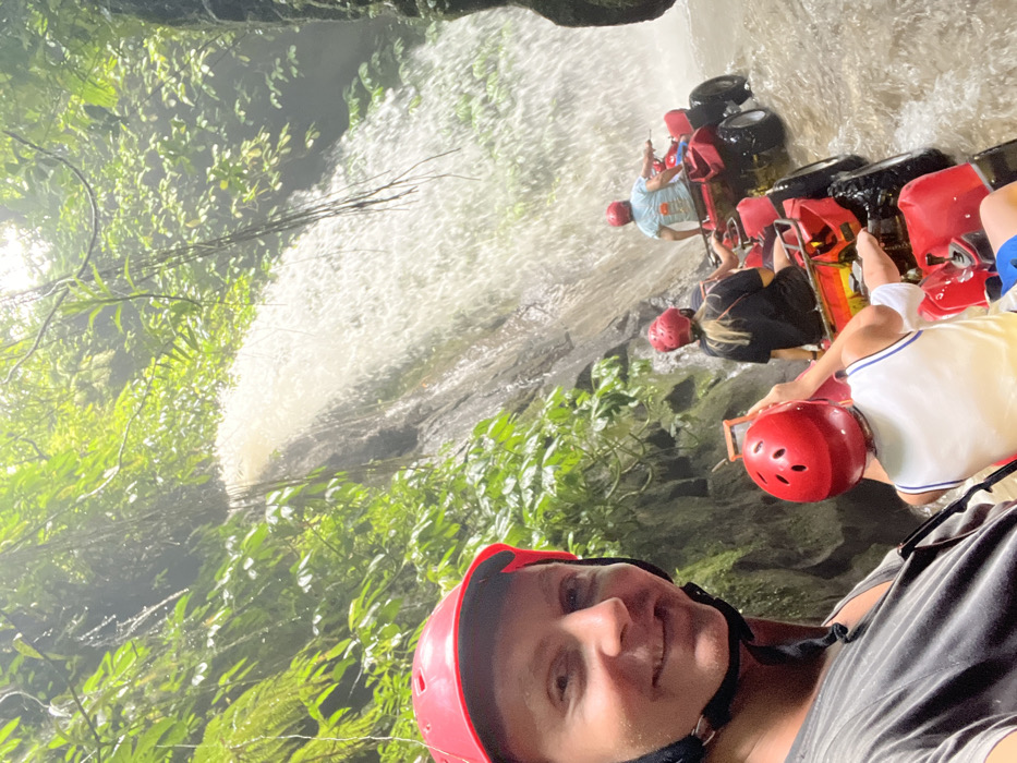 A selfie shows a smiling person wearing a red helmet. Behind them, other people are riding on the back of ATVs in front of a large waterfall. Lush green vegetation surrounds the waterfall and the ATV riders. The person in the foreground is wearing a gray t-shirt. The lighting suggests it might be daytime, but with dense foliage creating shade. The image is slightly rotated, making the scene appear diagonal.