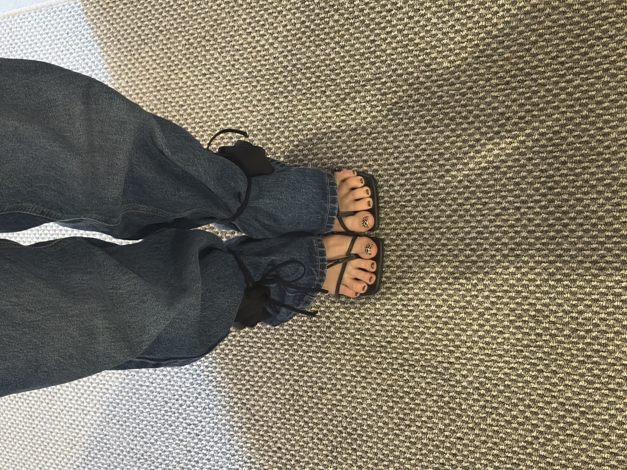 A person wearing denim jeans and black flat sandals stands on a patterned carpet. The jeans are tied with black fabric at the ankles. The person's toenails are painted dark brown, and the right big toe has a leopard print design. The scene takes place indoors, on a carpet that features a textured pattern of light and dark grey threads. The image does not provide any information about the city of Chaikovsky, Russia.