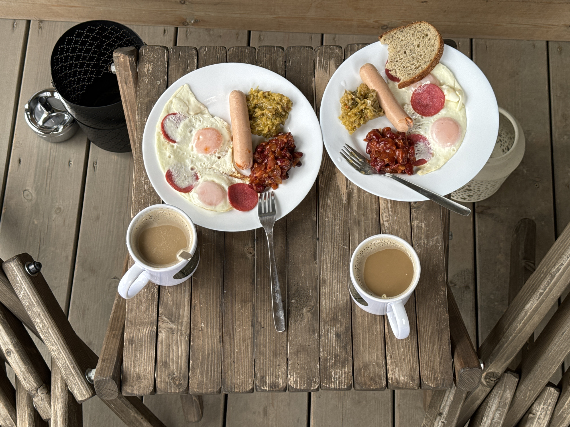 The image shows a rustic wooden table, likely outdoors, set for a meal. Two white plates each hold a breakfast of fried eggs, sausage, what appears to be sauerkraut, and a reddish-brown sauce. One plate includes a slice of bread. Two white mugs filled with coffee are also on the table. A fork sits between the plates. To the left of the table, there's a black metal object that could be a trash can or ashtray. The setting suggests a casual outdoor dining experience. There is no evidence in the photograph to support the location in Литвиново, Russia.