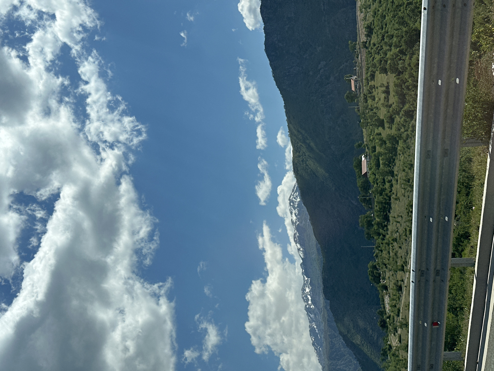 The image presents a mountainous landscape under a sky filled with scattered white clouds. A mountain range runs diagonally across the frame, with snow-capped peaks visible in the lower section. The slopes are densely covered with green vegetation.

To the right, a metal guardrail runs vertically, suggesting a roadside view. Beyond the guardrail, the land slopes towards the mountain. Small buildings or structures are visible amidst the greenery along the mountainside.

The image is likely taken from a moving vehicle on a road alongside the mountain. There are no people visible in the scene. Given the prompt's location, the image is likely taken in the vicinity of Klopot, Montenegro.