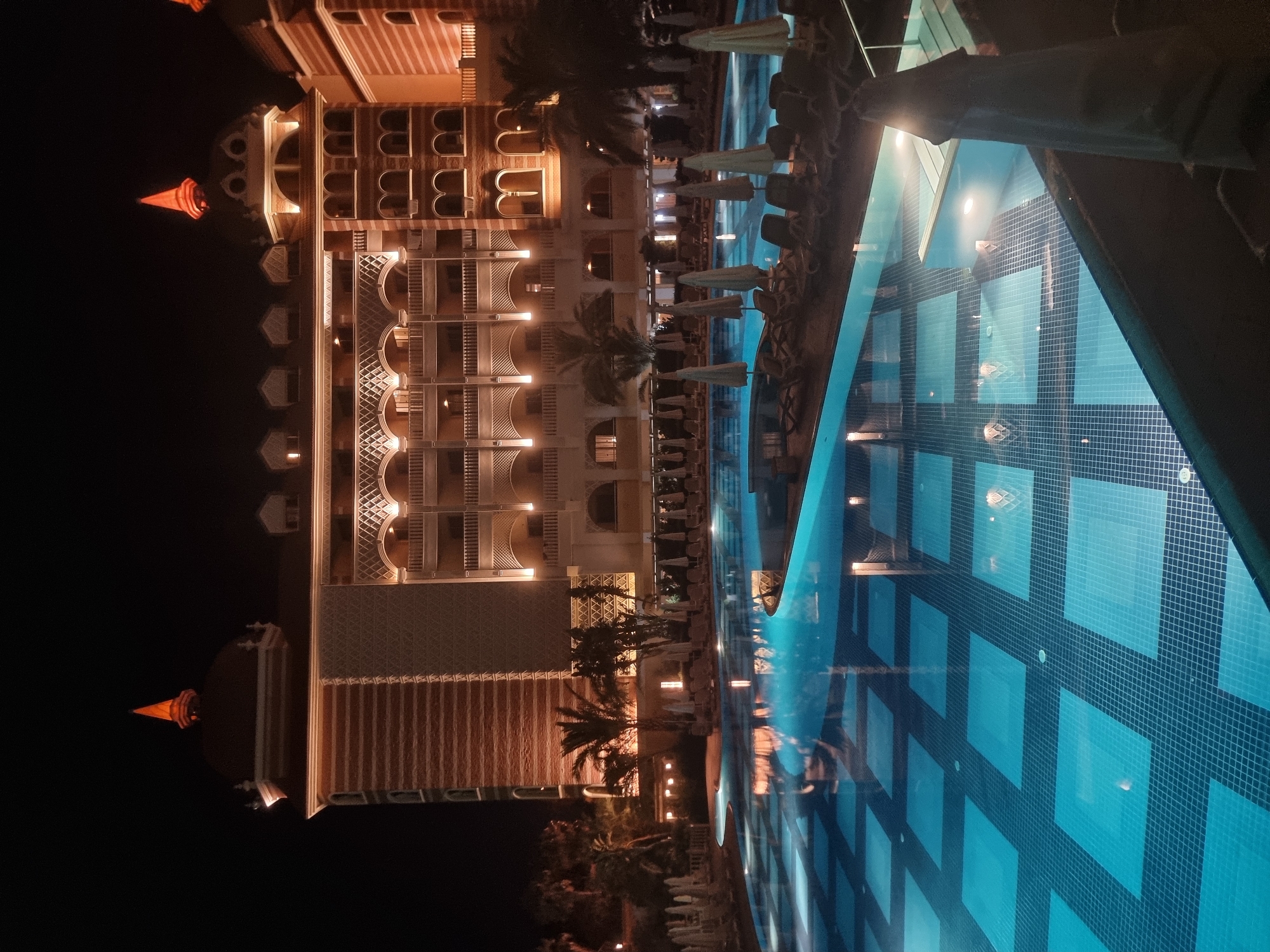 The image shows a resort at night in Manavgat, Turkey. A large, illuminated swimming pool with a tiled bottom is in the foreground, reflecting the lights of the building behind it. The building is a multi-story hotel with ornate architecture, featuring arched windows, balconies, and conical towers topped with orange lights. Palm trees and other tropical foliage are visible around the pool area, along with lounge chairs and closed umbrellas. The scene is lit by artificial lights, creating a warm and inviting atmosphere.