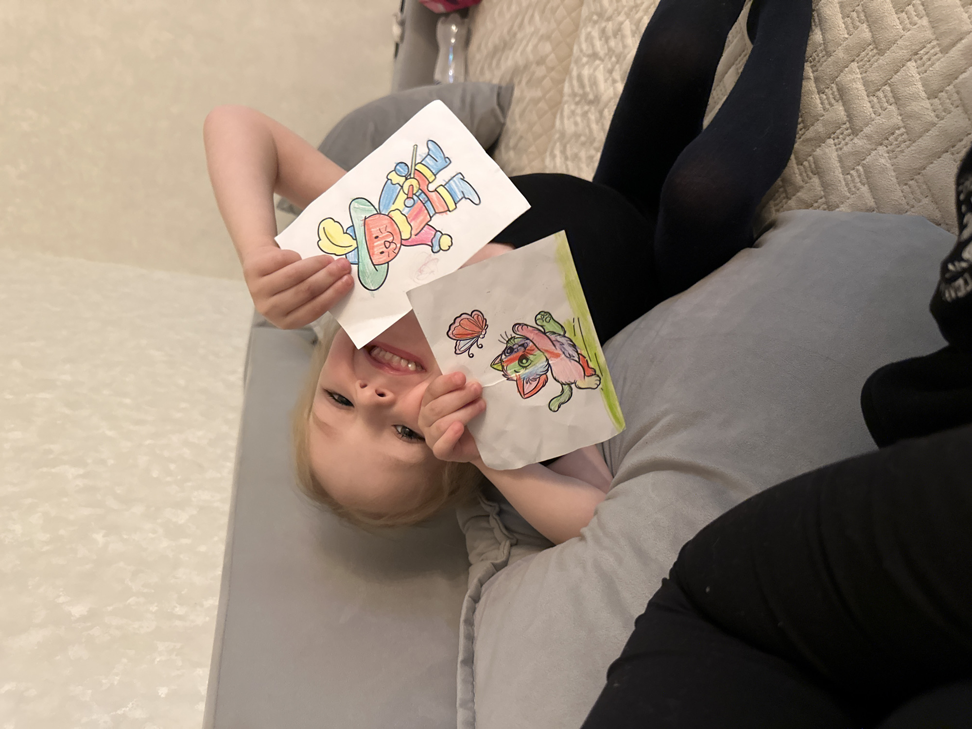 This indoor photograph features a young child, likely a girl with blonde hair, lying upside down on a comfortable light-colored sofa or bed, presenting her artwork. Her face is bright and joyful, illuminated by a wide smile that reveals her teeth.
She holds up two colorful drawings for the camera. In her left hand, she proudly displays a character, possibly a jester or musketeer, wearing a large hat, striped clothing, and boots, and holding a stick. In her right hand, closer to the foreground, is another vibrant drawing of a multi-colored cat-like creature with a butterfly hovering above it. Both pieces appear to be coloring pages that she has completed.
The child's legs, clad in black tights, are visible resting on the textured upholstery of the furniture, which might be a quilted pattern. Part of another person, possibly an adult or older child, also wearing black tights, can be seen to the right, suggesting they are sharing the seating. A grey cushion is visible beneath the child. The background is a simple, light-toned wall with a subtle, almost faded pattern. The scene exudes a sense of playful happiness and creative expression in a home setting in Chaikovsky, Russia. No discernible text is visible in the image.