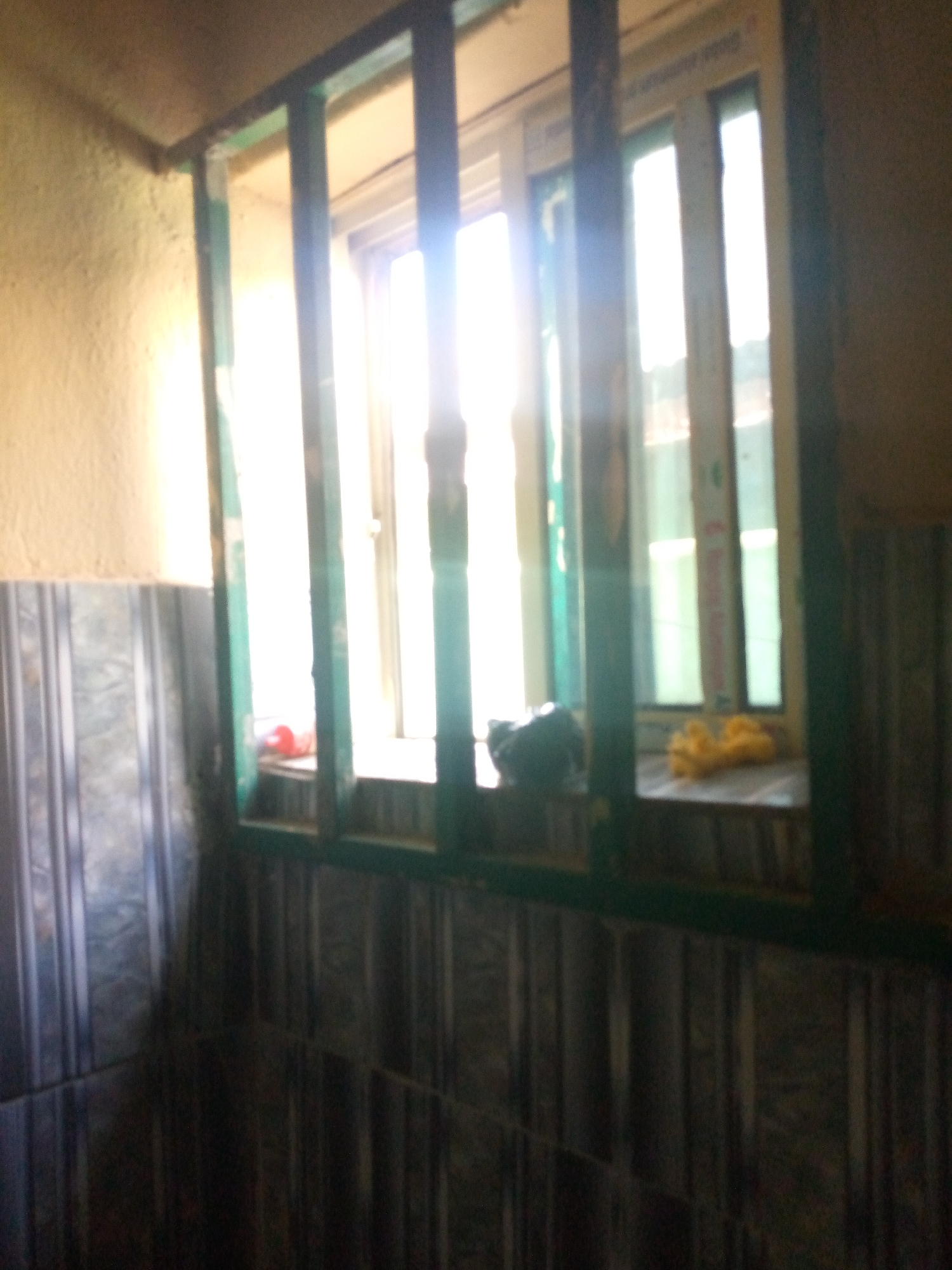 A slightly blurred, medium shot captures a corner of a room, focusing on a window with green bars. The window itself is divided into multiple panes, with bright daylight shining through, obscuring details outside. On the windowsill, a black bag is visible on the left, and a yellow object, possibly fabric, lies on the right. The wall to the left of the window has a textured, light-colored finish, while the wall below the window is tiled with a pattern of vertical grey and blue stripes. The floor area is dark and indistinct. The scene suggests an indoor space in Jalingo, Nigeria, possibly a residential or commercial building, with the window offering a view to the outside.