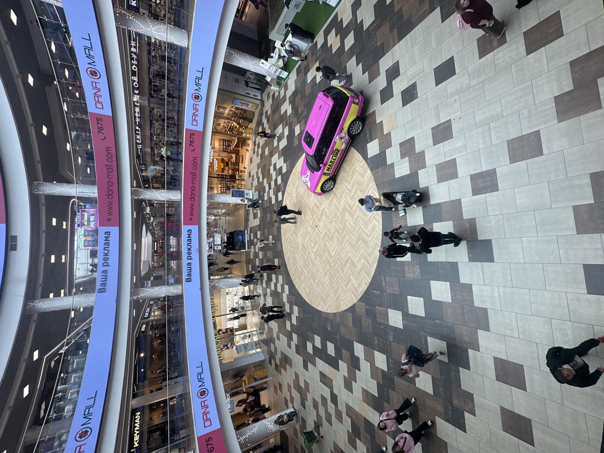 This image depicts the interior of a multi-level shopping mall, specifically Dana Mall in Minsk, Belarus. The scene is viewed from an elevated perspective, looking down onto the ground floor and the surrounding balconies. A prominent feature is the advertising screens that wrap around the upper levels of the mall, displaying "DANA MALL" and other promotional content.
On the ground floor, people are seen walking and interacting. A brightly colored pink and yellow SUV, branded with "MAXLINE" and images of cats, is parked in a central area. The floor is tiled in a patterned arrangement of various shades of brown, gray, and white. Shops and storefronts are visible on the different levels, including "L'OCCITANE." The overall environment is bright and spacious, characteristic of a modern shopping complex.