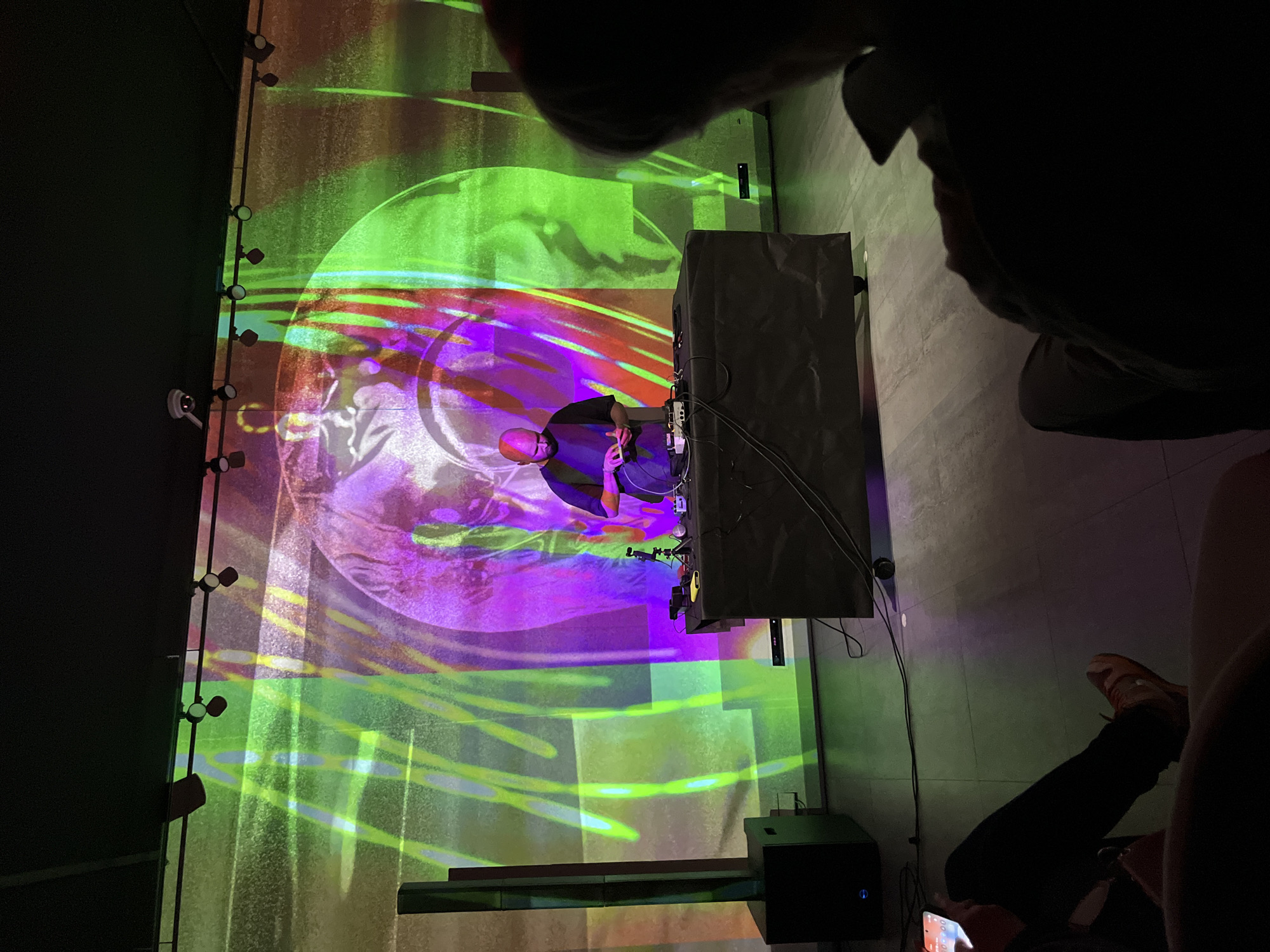 The scene is an indoor event in a city of Unknown, Unknown. A person with a shaved head is positioned in front of a DJ setup, consisting of electronic equipment housed in a black case. A colorful light display, with shades of green, purple, and red, is projected onto a wall behind the DJ. The wall also features a row of small lights along its edge. People are present in the foreground of the image, with only parts of their bodies visible, including a leg clad in black pants and tan shoes, and obscured facial features. A black speaker is positioned near the bottom of the frame. The environment appears to be a dimly lit space, with a tiled or concrete floor.