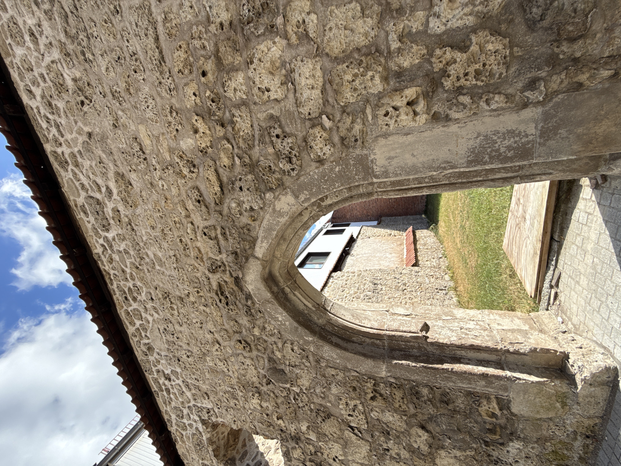 The image depicts a weathered, arched stone passageway in Walkenried, Germany, likely part of historical ruins. The rough-hewn, porous stone of the wall and archway contrasts with a small grassy area and a modern white building visible through the arch. The sky above is partly cloudy, with blue skies and white clouds. The ground in the foreground is paved with stone tiles. There are no people or animals in the scene.