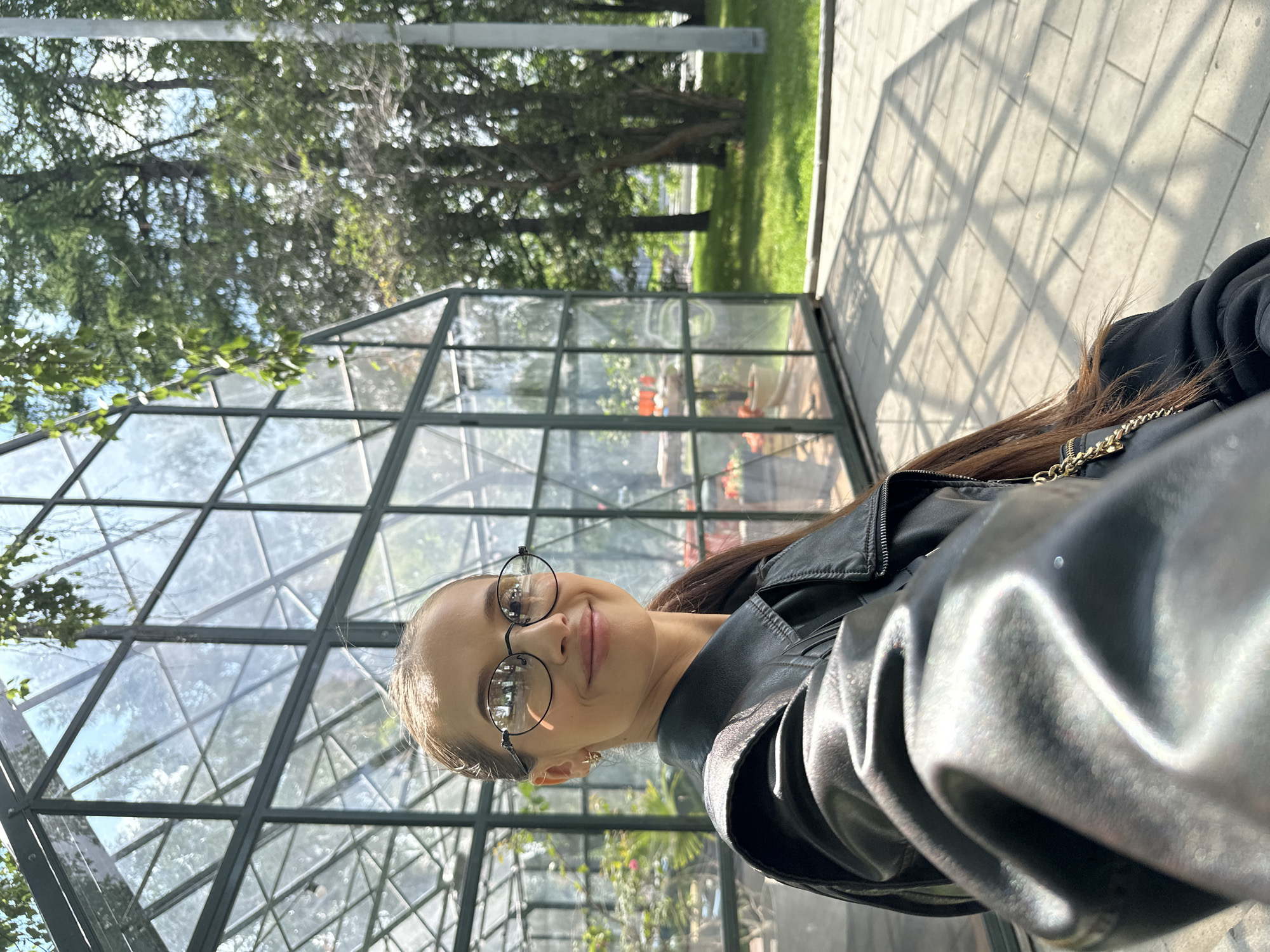 A young woman with long brown hair and glasses is taking a selfie in front of a greenhouse. She is wearing a black leather jacket. The greenhouse has a green metal frame and glass panels, and it appears to be located in an outdoor park or botanical garden. Trees and grass are visible in the background. The ground is paved with light-colored tiles, and shadows from the greenhouse structure are cast across the pavement. The overall scene suggests a bright, sunny day. The location is identified as the Yekaterinburg City Park of Culture and Rest, located at 18 Mayakovsky Street, Yekaterinburg, Russia.
