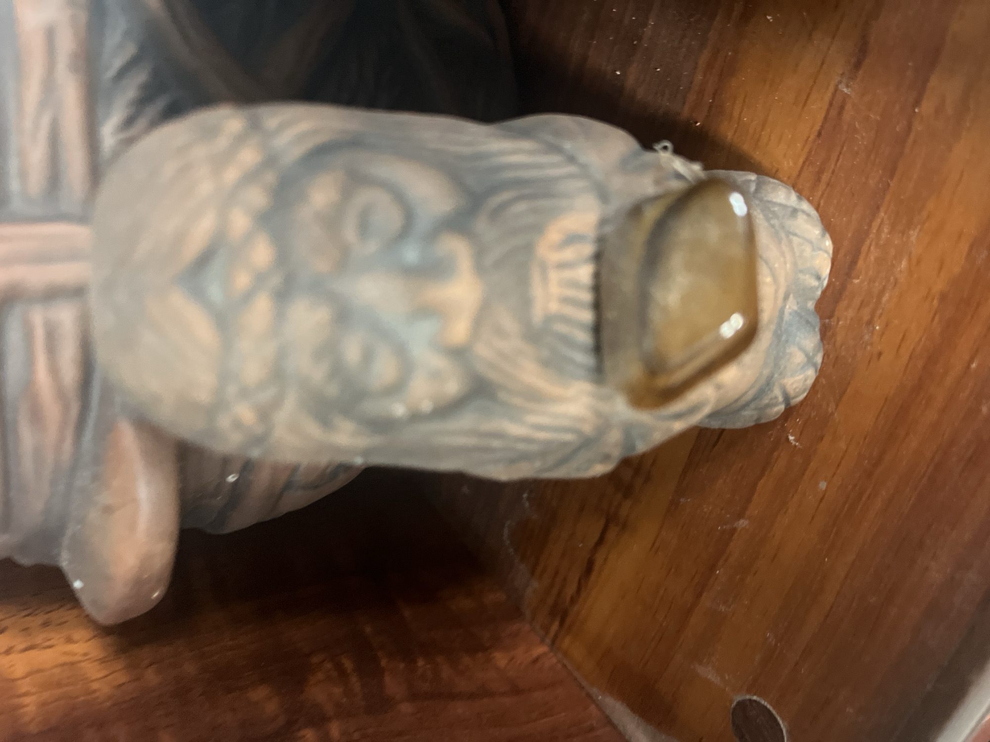 The image shows a close-up view of a carved wooden or clay figurine with a beard and a crown-like headdress. The figurine's face is detailed with carved eyes, nose, and mouth. A smooth, polished stone or a piece of amber is embedded in the figurine's mouth area. The figurine appears to be part of a larger, unfinished religious or decorative piece, with glimpses of brown material forming a background. The surface it rests on is a polished wooden surface with a visible grain. There is no information about the location in Vladimirovka, Russia, in the image.