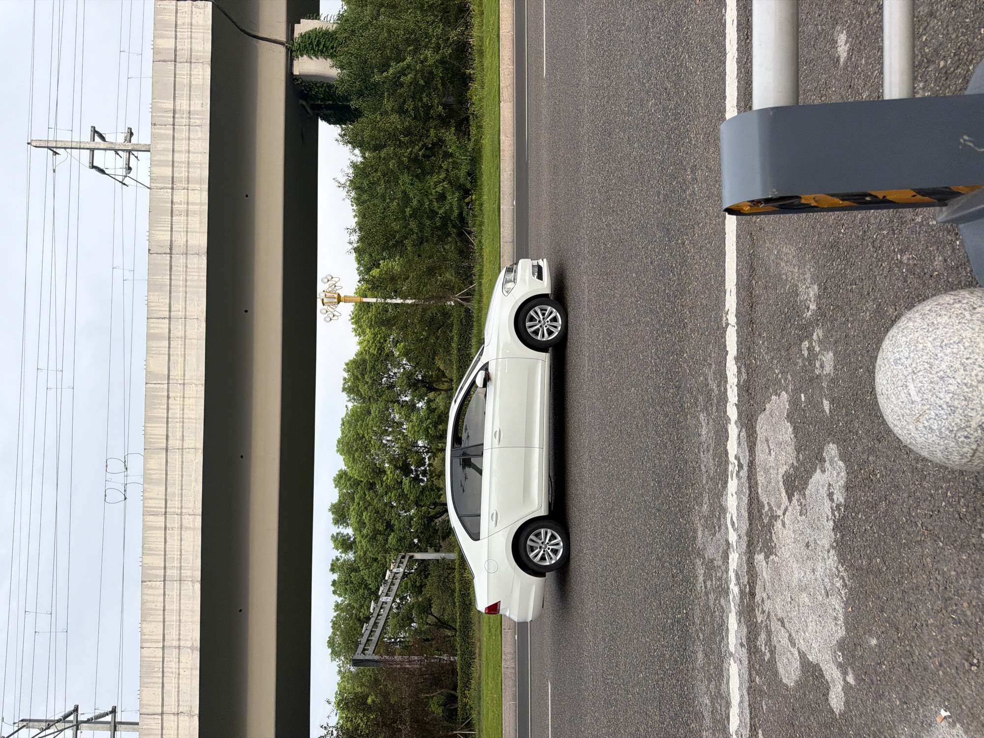 The image captures a white sedan driving on a multi-lane road in Jiangjin, China. The car is positioned in the middle lane, with its left side facing the camera. The road surface appears to be asphalt, with visible white lane markings. In the foreground, a portion of a grey barrier with yellow and black warning stripes is visible on the right, and a speckled grey spherical object is on the lower left.
Above the road, a concrete overpass or bridge spans across the scene, supported by columns that are partially obscured by lush green trees. The sky is overcast. Overhead power lines run parallel to the overpass. The background is filled with dense foliage and trees, suggesting a semi-urban or suburban setting. A street lamp with multiple bulbs is also visible behind the car. The overall impression is that of a transit area within a city, likely with elevated infrastructure.