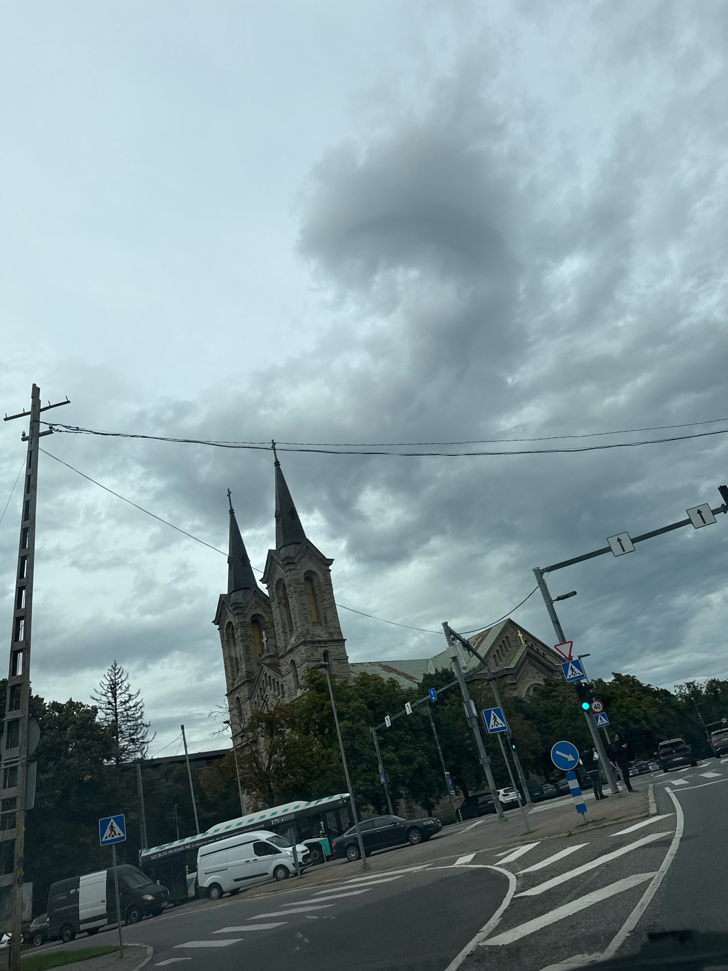 In Tallinn, Estonia, a cloudy day is depicted with a view of the St. Peter's Church, an iconic Lutheran church with two prominent spires. The foreground shows a street scene with a traffic light, various vehicles including a bus, vans, and cars, and pedestrian crossing signs. A wooden utility pole with wires runs diagonally across the frame, partially obscuring the sky and the church. The church's stone facade and architectural details are visible against the overcast sky. The image captures a street intersection with road markings and street signs, suggesting an urban setting. The overall atmosphere is muted due to the cloudy weather.