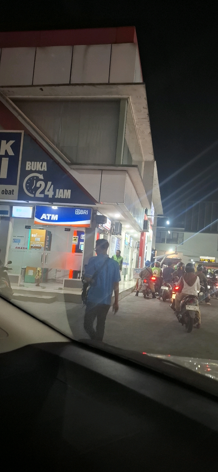 The image depicts an outdoor scene at night, likely in Surakarta, Indonesia, showing a brightly lit convenience store or mini-market attached to a gas station. The store features a prominent sign that reads "BUKA 24 JAM" (Open 24 Hours) with a clock graphic. Below this, there is an ATM sign for BRI (Bank Rakyat Indonesia). Several ATMs are visible through the glass storefront, and the interior is illuminated.
In front of the store, a man in a blue shirt walks away from the camera, carrying a dark bag over his shoulder. To the right of the store, a queue of motorcycles with riders is waiting, suggesting activity at the gas station or store. Some of the motorcycles have their headlights on, casting light on the pavement. The overall environment appears to be an urban setting at night, with artificial lighting dominating the scene. The perspective is from inside a vehicle, looking out through the windshield.