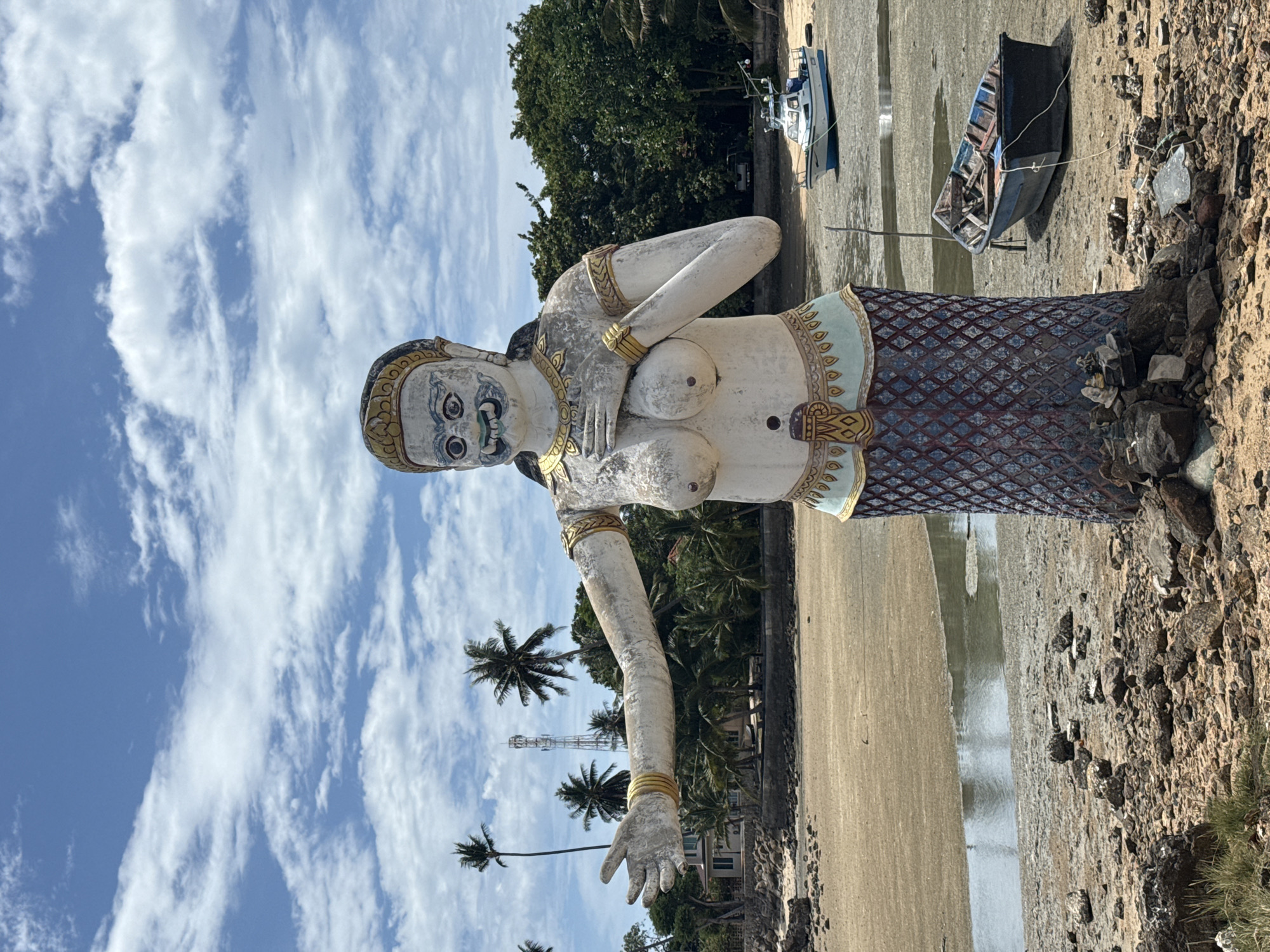 A full-shot, eye-level, outdoor, daytime photograph captures a weathered, stucco statue of a mythical female creature positioned on a rocky, sandy shore. The statue is predominantly white with blue and gold accents. It features a human-like upper body with prominent breasts, an ornate gold headpiece, and a lower body resembling a scaled skirt. One arm is outstretched to the viewer's left, while the other rests on its chest.
The statue is situated on a beach that appears to be at low tide, with a small body of water visible in the mid-ground. Beyond the statue, a sandy expanse stretches towards a line of lush green trees and palm trees. In the distance, a modern telecommunications tower is visible amongst the foliage. Two boats are present on the shore: a small, white, motorboat with a blue hull in the mid-ground, and a derelict, blue wooden rowboat filled with debris in the foreground. The sky above is a vibrant blue, dotted with fluffy white clouds.
The scene is located in the city of Ko Samui District, Thailand, specifically on the beach of Ban Tai, near the local pier.