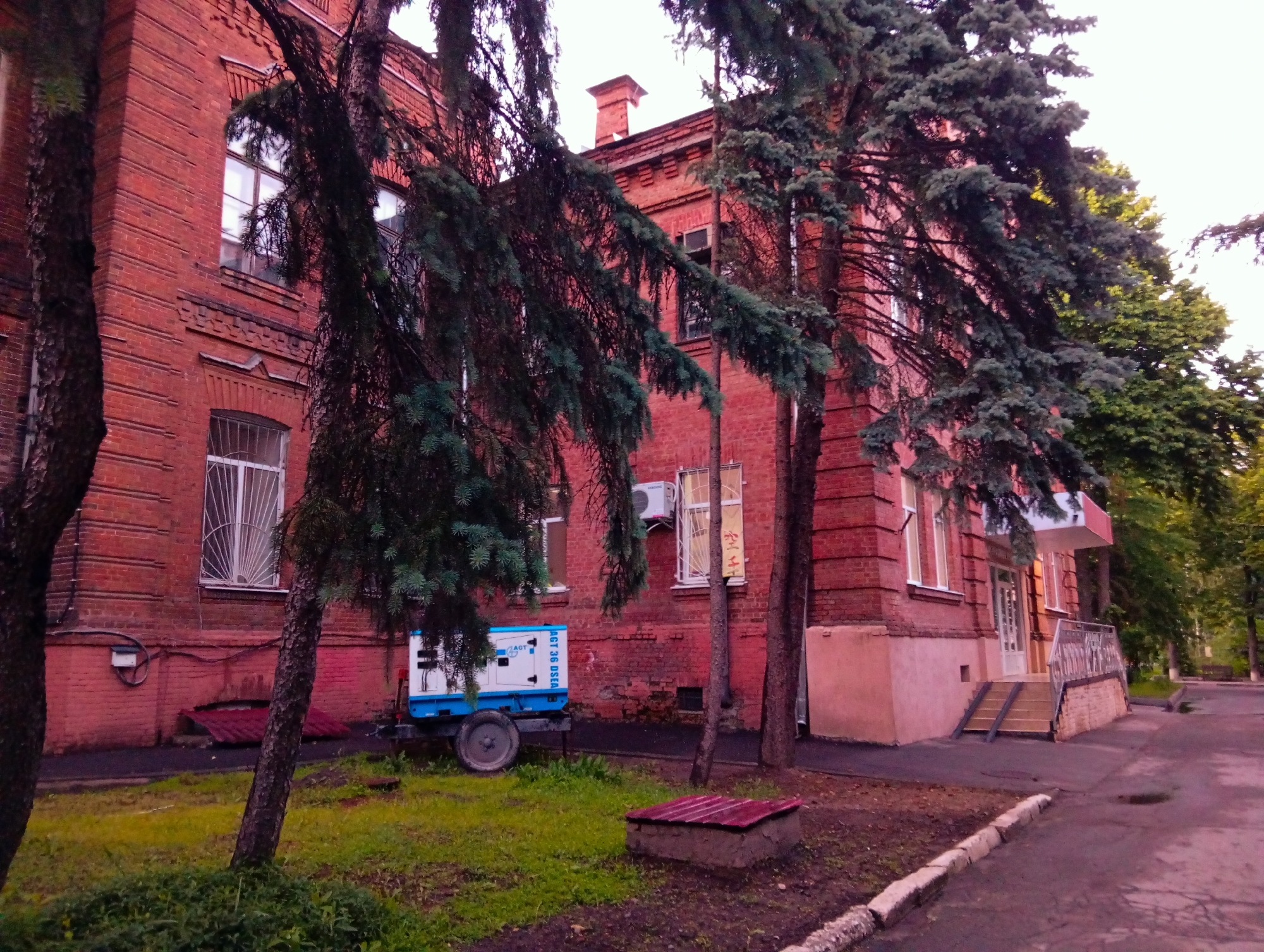 The image shows a red brick building partially obscured by trees, likely taken on a street in Poltava, Ukraine. The building appears to be a two-story structure with a protruding entrance on the right side, accessed by a set of steps with metal railings. A blue and white trailer or generator unit is parked on the grass in front of the building. The foreground includes grass and some ground cover, with a section of sidewalk visible on the right. The sky is overcast. No people are visible in the image.
