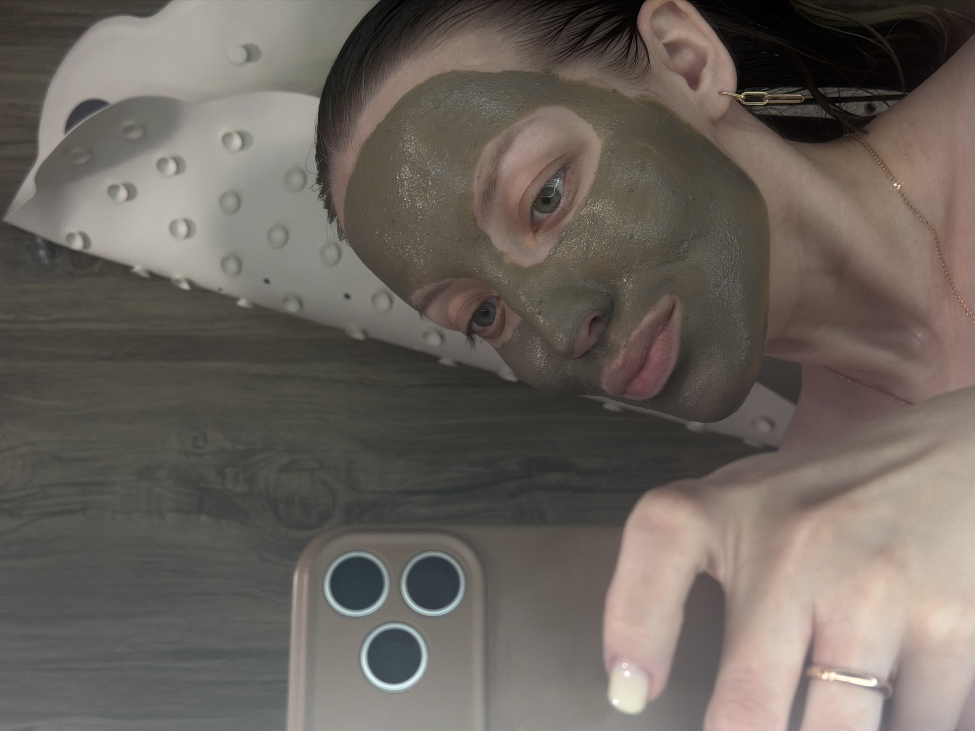 A woman with a greyish-green facial mask is lying down, taking a selfie with a gold-colored smartphone. She is on a white, textured surface that resembles a bath pillow. Her hair is wet and slicked back. She wears gold link earrings and a delicate gold necklace. She has a simple gold band ring on her finger. The background appears to be a dark wood-grain surface. The location is unknown; there is no information in the image to indicate it is Reshetnikovo, Russia.