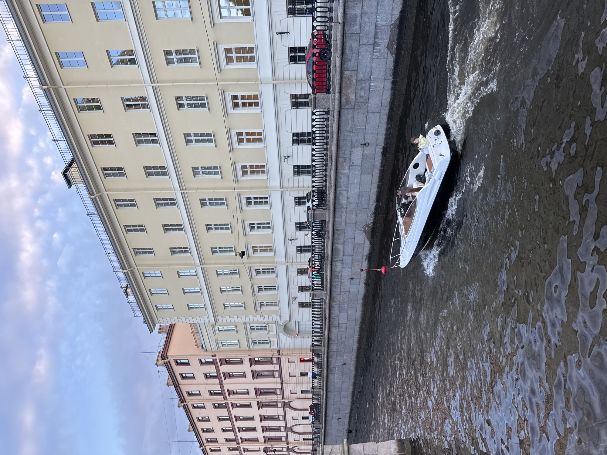 A motorboat with three passengers navigates a canal in Saint Petersburg, Russia. The boat, white with a dark hull, is moving at speed, leaving a wake. The passengers, two adults and a child, are seated within the vessel. On the canal's embankment, a row of buildings lines the waterfront. The buildings are multi-story structures with numerous windows. The architecture suggests a historic European city. A black metal railing with ornate detailing runs along the top of the embankment, separating the canal from the street and buildings. Cars are parked along the street behind the railing. The sky above is overcast with clouds. The water in the canal is dark and appears slightly rippled, reflecting the buildings and sky. A small red buoy with a white top floats in the water near the boat. The scene depicts a typical urban canal environment in Saint Petersburg.
