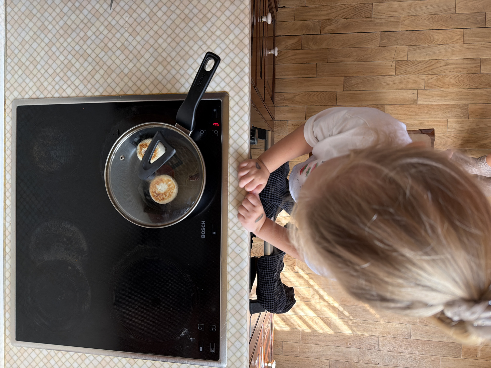 An overhead, high-angle shot shows a child standing in front of a black induction cooktop with two small pancakes frying in a pan. The cooktop has several control buttons and a digital display showing "3". The pan has a glass lid and a black handle, and a black spatula is visible inside, resting on one of the pancakes. The countertop is tiled with a beige and white diamond pattern. The child, seen from behind, has blonde hair tied in a ponytail and is wearing a white shirt. Their hands, which are resting on the edge of the counter, have some markings on them. The floor is made of light-colored wooden planks, with diagonal shadows cast by sunlight. The scene appears to be in a domestic kitchen in Khimki, Russia.