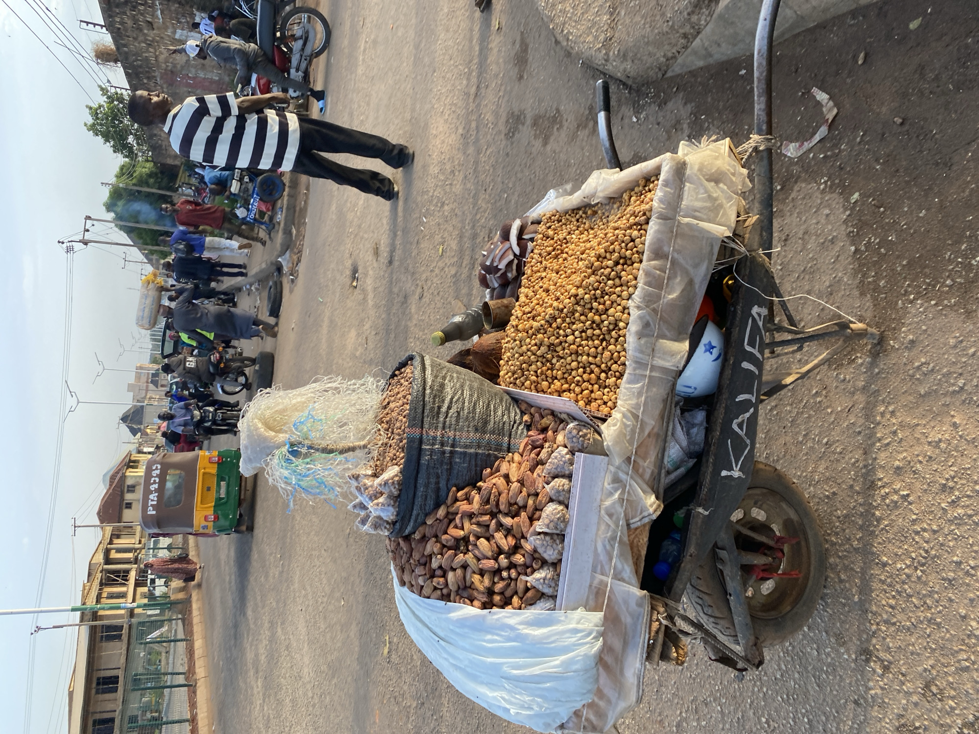A street scene in Bukuru, Nigeria, depicts a vendor's cart filled with various food items. The cart, resting on a single front wheel, is laden with peanuts, dates, and unidentified objects in bags and containers. The name "KALI" is written on the side of the cart. A man in a striped shirt and black pants stands beside the cart, appearing to observe the scene. In the background, there is a crowd of people and motorcycles on the road, along with a yellow and green tricycle taxi bearing the registration number PTA-4345. Buildings and utility poles line the street. The scene suggests an active marketplace or roadside vending area.