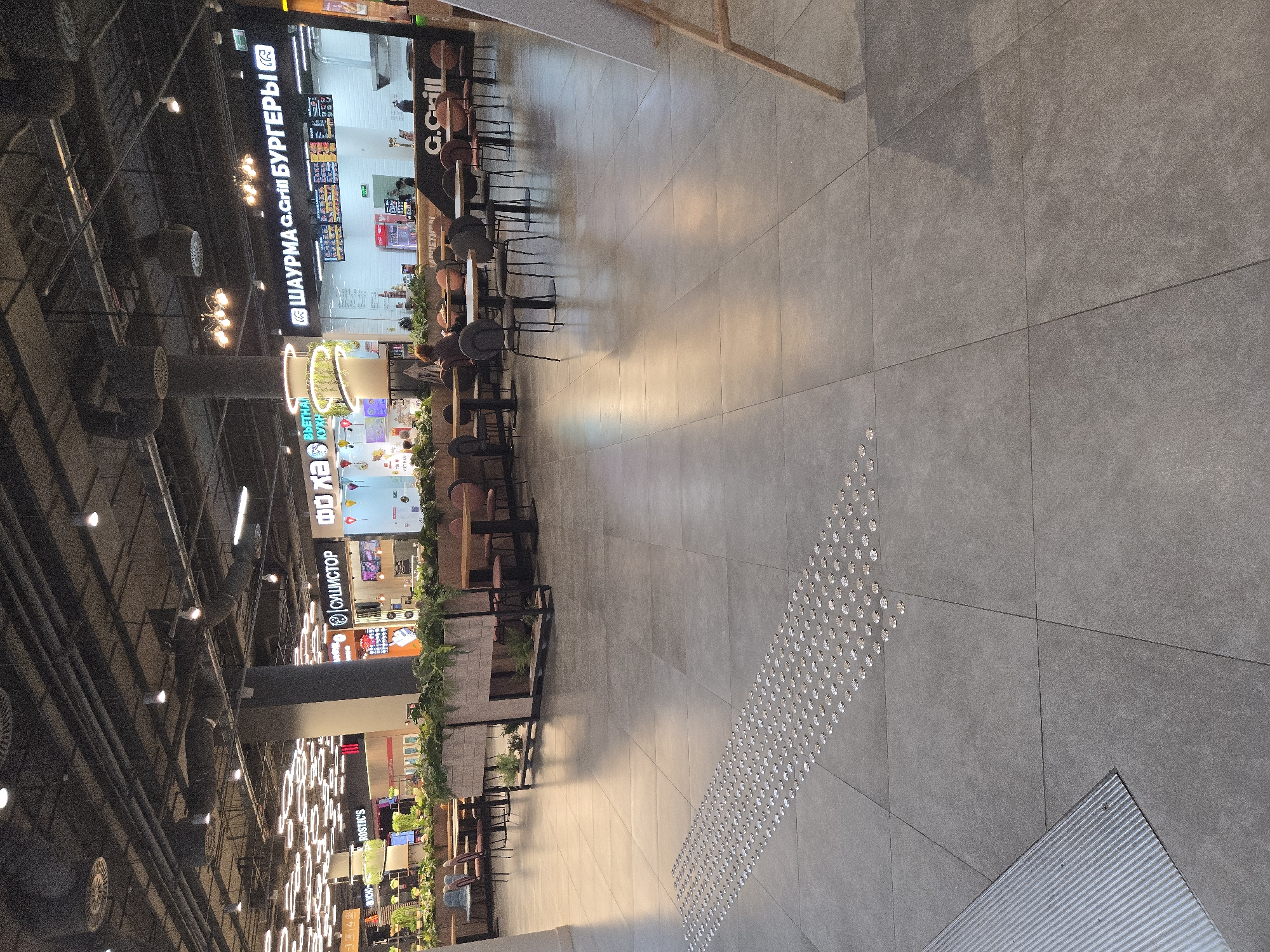The image shows a wide, well-lit indoor space, likely a shopping mall or food court, in Moscow, Russia. The floor is tiled with large, gray tiles, and a row of tactile paving stones guides the way diagonally across the frame.
On the left side of the image, a row of food stalls and shops is visible. One prominent sign reads "ШАУРМА G.Grill БУРГЕРЫ" in large white lettering against a dark background, indicating a food establishment. Further down, another sign displays "ФОХА" in large, stylized letters, and below that, "СУШИСТОP" with a red logo. These signs suggest various food options are available. The stalls have modern interiors with bright lighting and display menus. Greenery, in the form of potted plants and hedges, is strategically placed between the food outlets and seating areas, adding a touch of nature to the urban setting.
Several tables and chairs are set up in front of the food stalls, with some patrons seated and eating. The lighting in the scene is a mix of overhead fluorescent lights and decorative circular light fixtures on the ceiling, suggesting it is daytime or evening within the enclosed space. The overall atmosphere appears to be calm and ordinary, with no specific event taking place beyond the usual commercial activity of a mall. The building's infrastructure, including pipes and ventilation systems, is visible on the ceiling in some areas, hinting at the functional aspects of the setting.