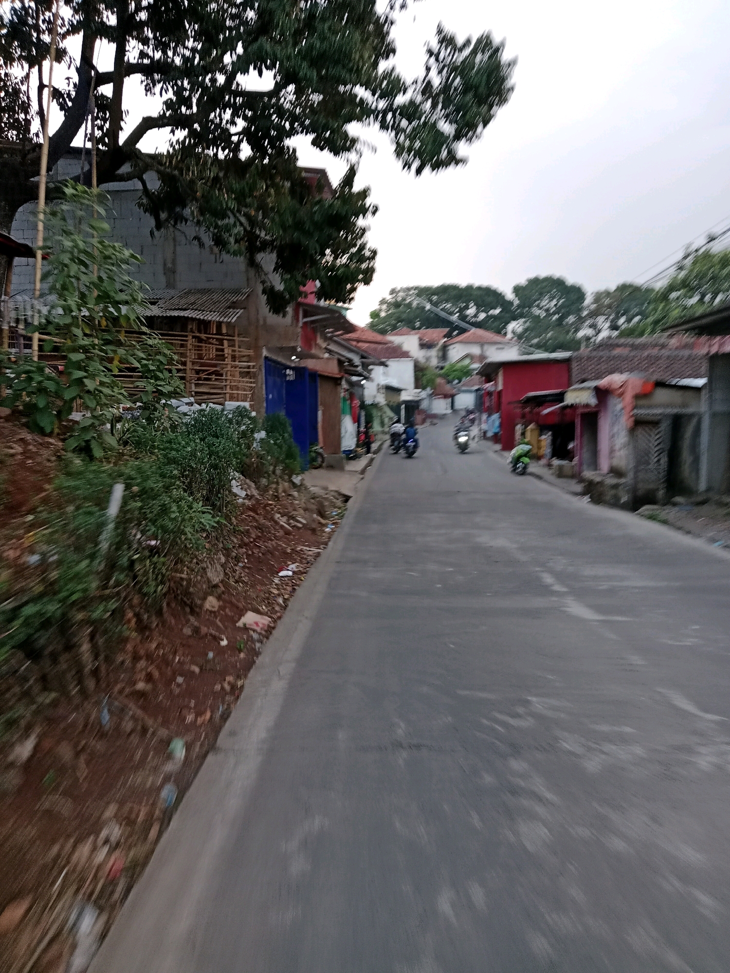 The image shows a street scene in Cilaku, Indonesia. On the left side of the street, there is a steep embankment covered in dirt and vegetation. The embankment has some debris on it, including plastic bottles and bags. Beside the embankment, there is a narrow paved road. On the right side of the road, there are several small buildings, some of which appear to be houses or shops. The buildings have corrugated metal roofs and some have red paint. There are a few motorcycles on the road, and one is parked near a building. The sky is overcast, and the overall impression is one of a modest, somewhat rural urban environment. The presence of trees and vegetation suggests a tropical climate.