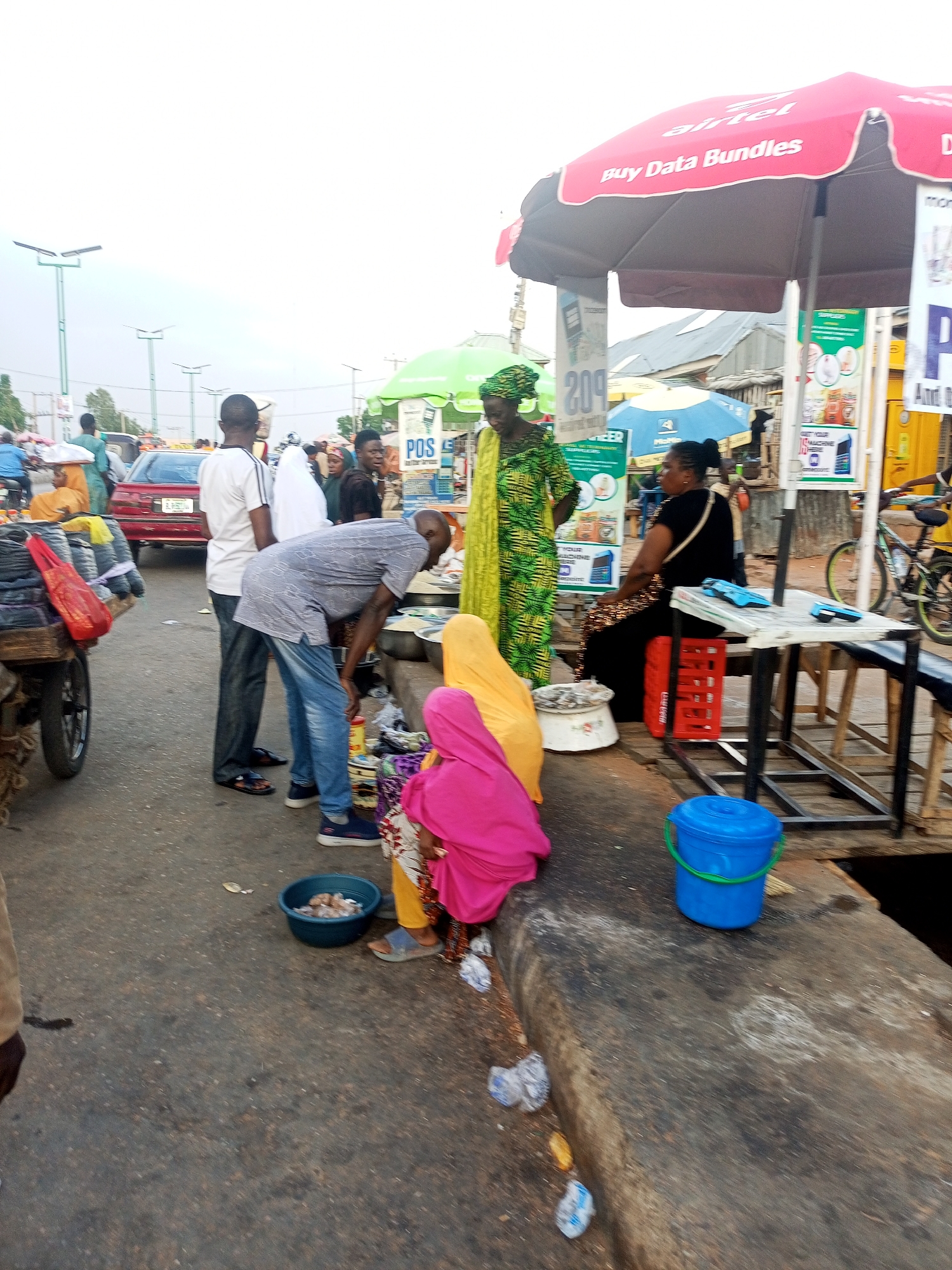 The image depicts a bustling market scene in Gombe, Nigeria. A group of people are gathered around a vendor selling goods, possibly food items, displayed in bowls on a concrete surface. Several individuals are examining the merchandise, while others stand nearby. A woman in traditional attire stands behind the vendor. A red car is parked nearby, and an Airtel umbrella offers shade to a vendor sitting nearby. A cart loaded with goods is partially visible. There are lampposts and other buildings visible in the background indicating an urban setting.
