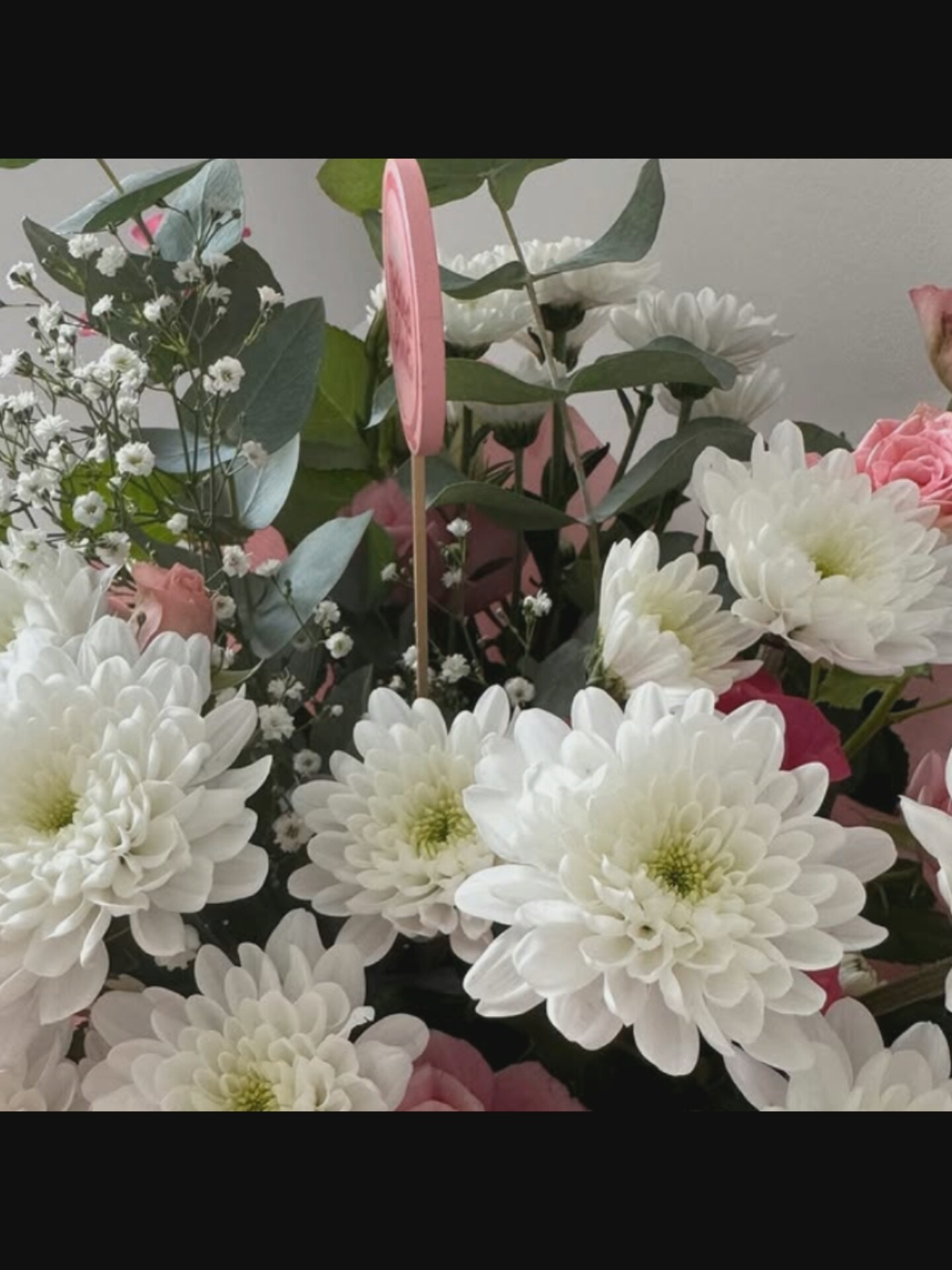 A close-up shot captures a bouquet of flowers, predominantly white chrysanthemums with some delicate pink roses and wisps of baby's breath interspersed. Green eucalyptus leaves add a touch of foliage. A pink circular party favor is also visible, sticking out from the arrangement. The background is a plain, light-colored wall. There is no information to suggest the image is from Bucharest, Romania.