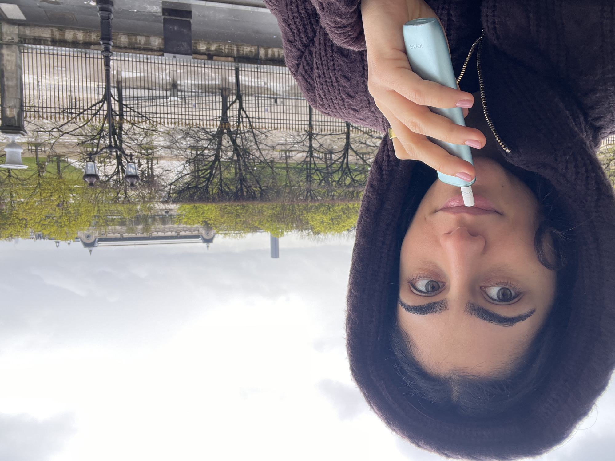 A female individual is depicted in a close-up, facing the viewer from the left side of the frame, with her hood pulled up. She has dark hair, prominent eyebrows, and brown eyes. Her right hand, with light pink painted nails and a gold ring on the ring finger, holds a light blue and white IQOS heated tobacco device positioned near her mouth. She is wearing a dark brown, textured, zippered hooded garment.
The background reveals an urban landscape under an overcast sky. A dark wrought-iron fence with decorative accents runs horizontally across the mid-ground. Beyond the fence, an open area with bare trees and trees with fresh green foliage is visible, appearing wet, suggesting recent rain. To the right of the frame in the mid-ground, a black, ornate Parisian streetlamp with two lanterns stands. A classical white stone urn on a pedestal is also visible on the right.
In the distant background, across the Seine River in Paris, France, the distinctive Beaux-Arts architecture of the Musée d'Orsay, characterized by its long structure and prominent domed roofs, is clearly visible. Further to the left and behind the Musée d'Orsay, the tall, slender profile of the Tour Montparnasse skyscraper is discernible. The scene suggests a vantage point from the north bank of the Seine, possibly near the Tuileries Garden, looking south towards the museum on the Rive Gauche.