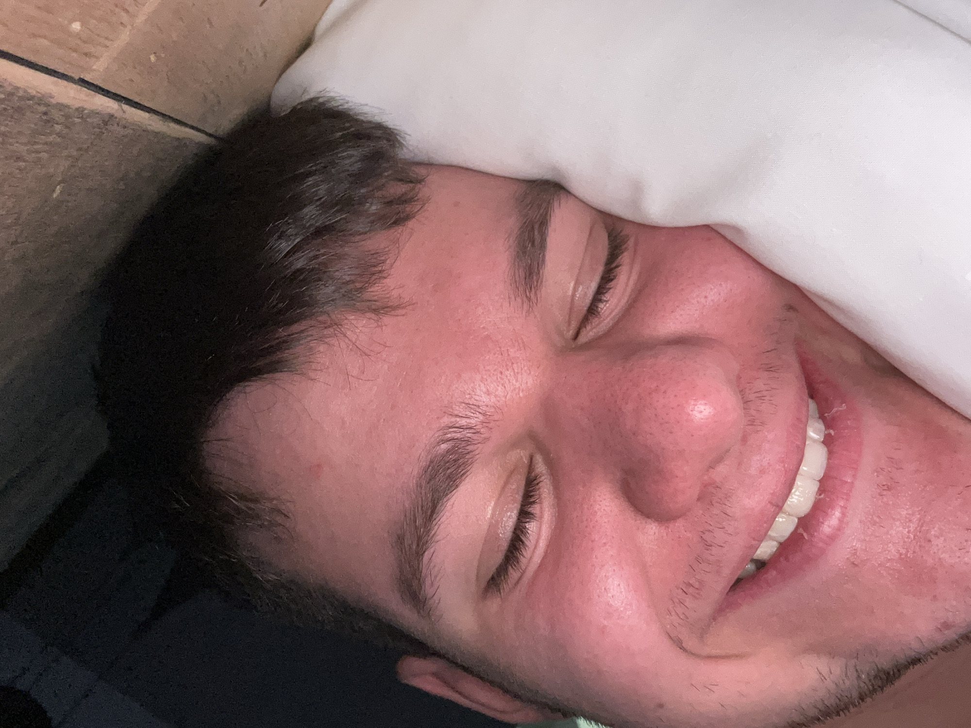 A close-up image shows a man lying in bed, appearing relaxed or asleep with a slight smile. He is positioned on his side, with a white pillow partially covering his forehead. His eyes are closed, and his dark hair is tousled. The man has fair skin, dark eyebrows, and stubble. To the upper left, there is a wooden surface. The location, Pamporovo, Bulgaria, is not discernable from the image.