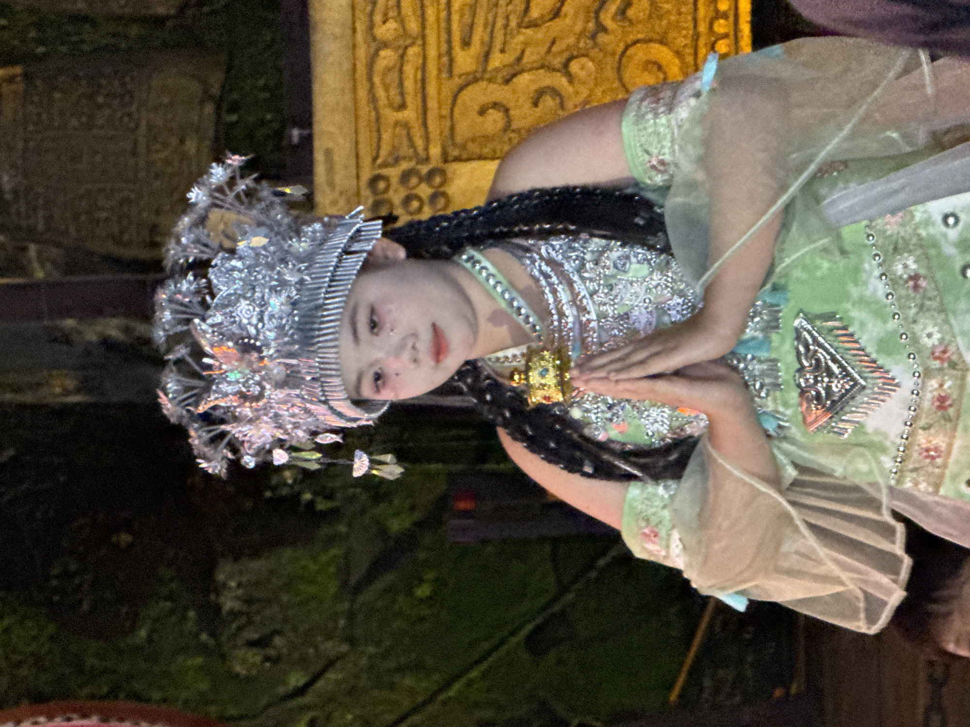 A woman in traditional attire is captured in an image taken in the Yuzhong District, China. She is adorned with an elaborate silver headdress. Her clothing includes a heavily decorated green dress, with silver adornments, layered necklaces, and sheer sleeves. Her hands are clasped together in front of her. The background is dark, with hints of foliage, and a carved yellow stone structure or pillar.