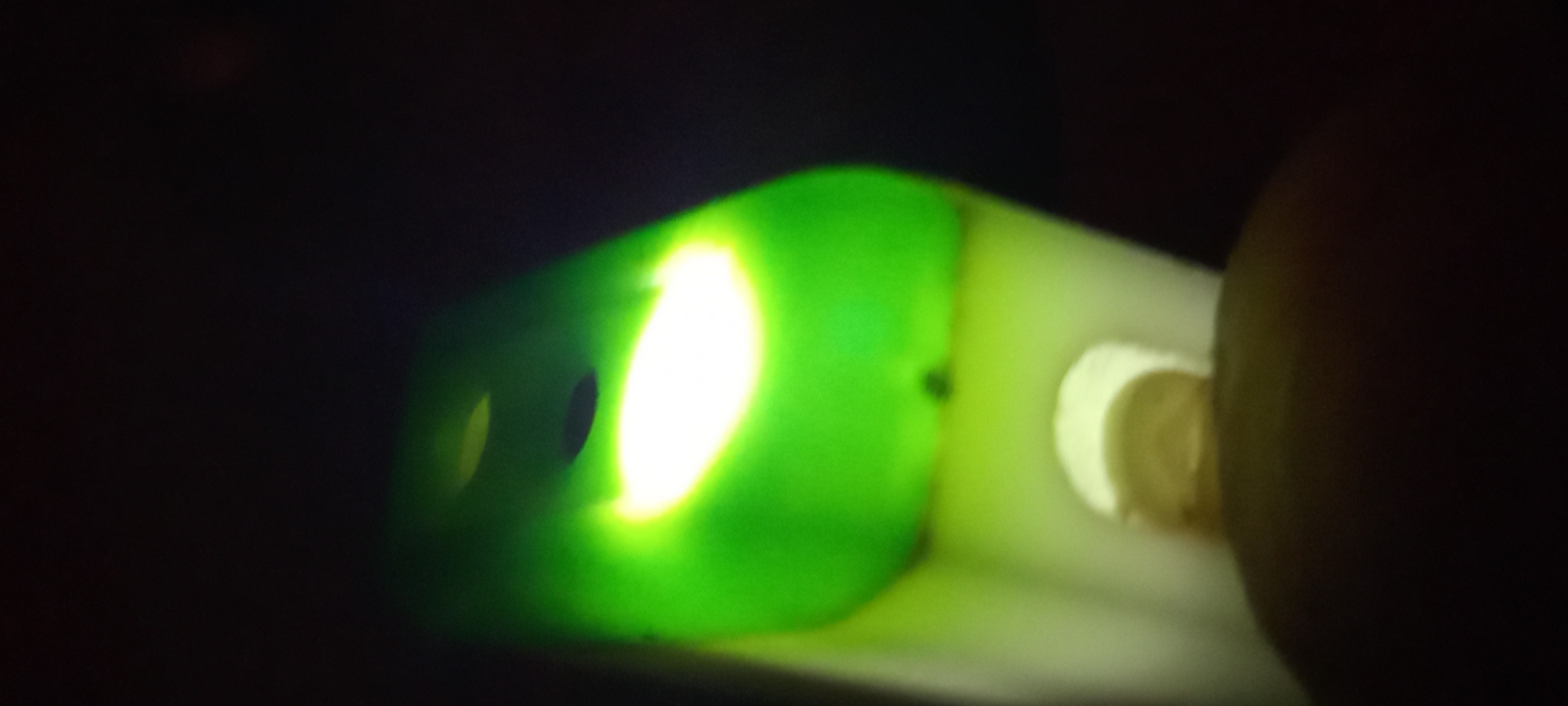 The image displays a close-up, blurred view of a green, translucent object with a bright light source shining through it, creating a halo effect. Two dark, oval-shaped openings are visible on the surface of the object. Below the green object, a lighter, cylindrical structure extends downwards, and at the very bottom, a dark, rounded mass is partially visible. The background is entirely black, suggesting a dark environment or the absence of any discernible background details. The image lacks any identifiable landmarks or contextual clues that would allow for its location to be determined in Kryvyi Rih, Ukraine, or any other city.
