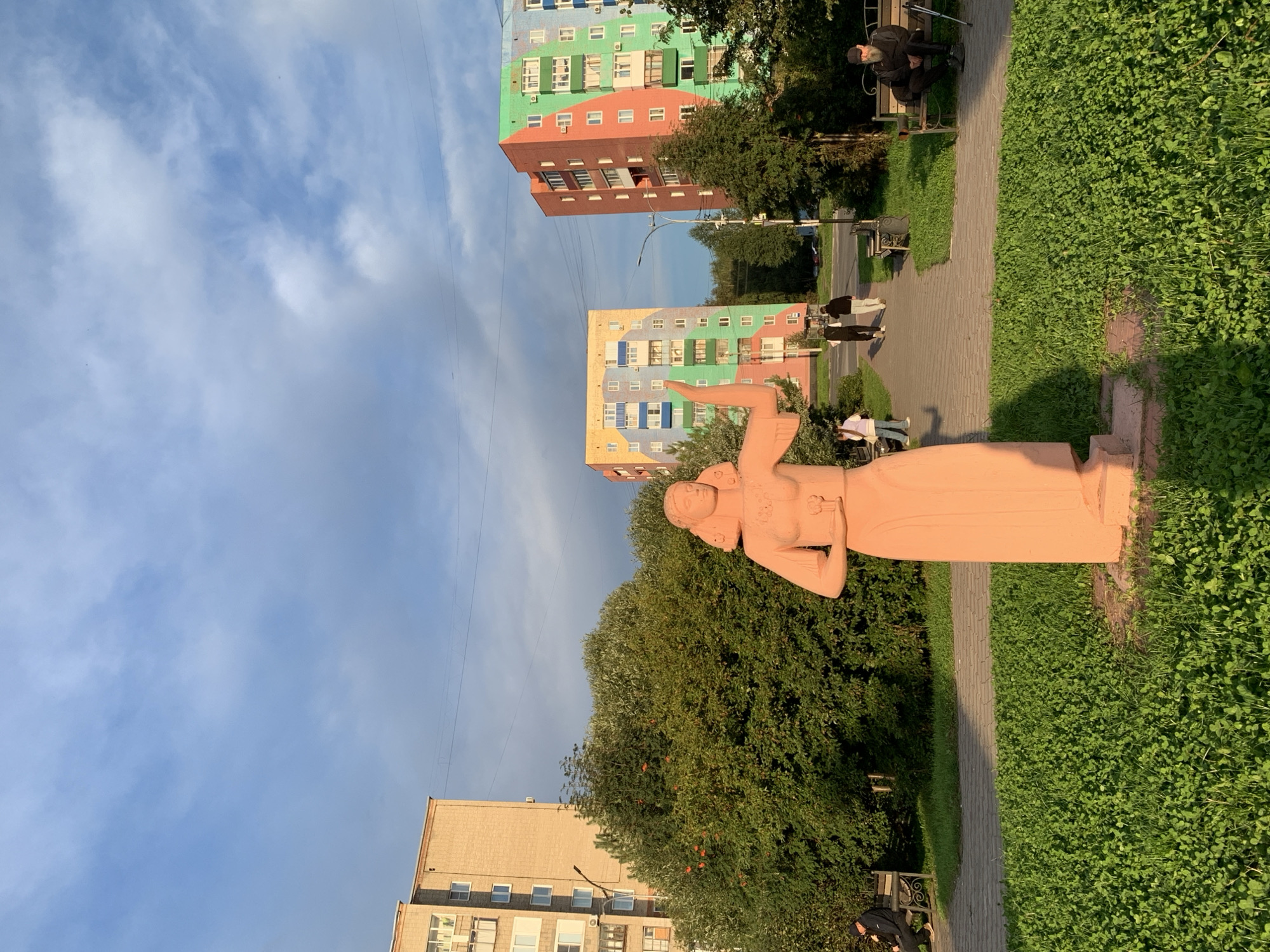 A statue of a woman, sculpted from light pink material and standing tall, is positioned in the foreground of the image. She is depicted wearing a long, flowing dress and holding a small object in her outstretched hand. Behind her, a patch of vibrant green grass is visible, with a paved walkway extending into the middle ground. Several people are present in the scene: one individual is seated on a bench to the left, another is walking on the path with two companions, and a man is seated on a bench to the right, accompanied by walking sticks. The background is dominated by residential buildings, with a tall, tan-colored apartment block to the left and brightly colored, multi-story apartment buildings to the right, adorned with red, green, and yellow facades. Lush green trees, including a large bushy tree to the left of the statue, add to the urban landscape. The sky above is a mix of blue and white clouds, illuminated by what appears to be the warm glow of either sunrise or sunset, casting long shadows. The scene is set in the city of Prokopyevsk, Russia.