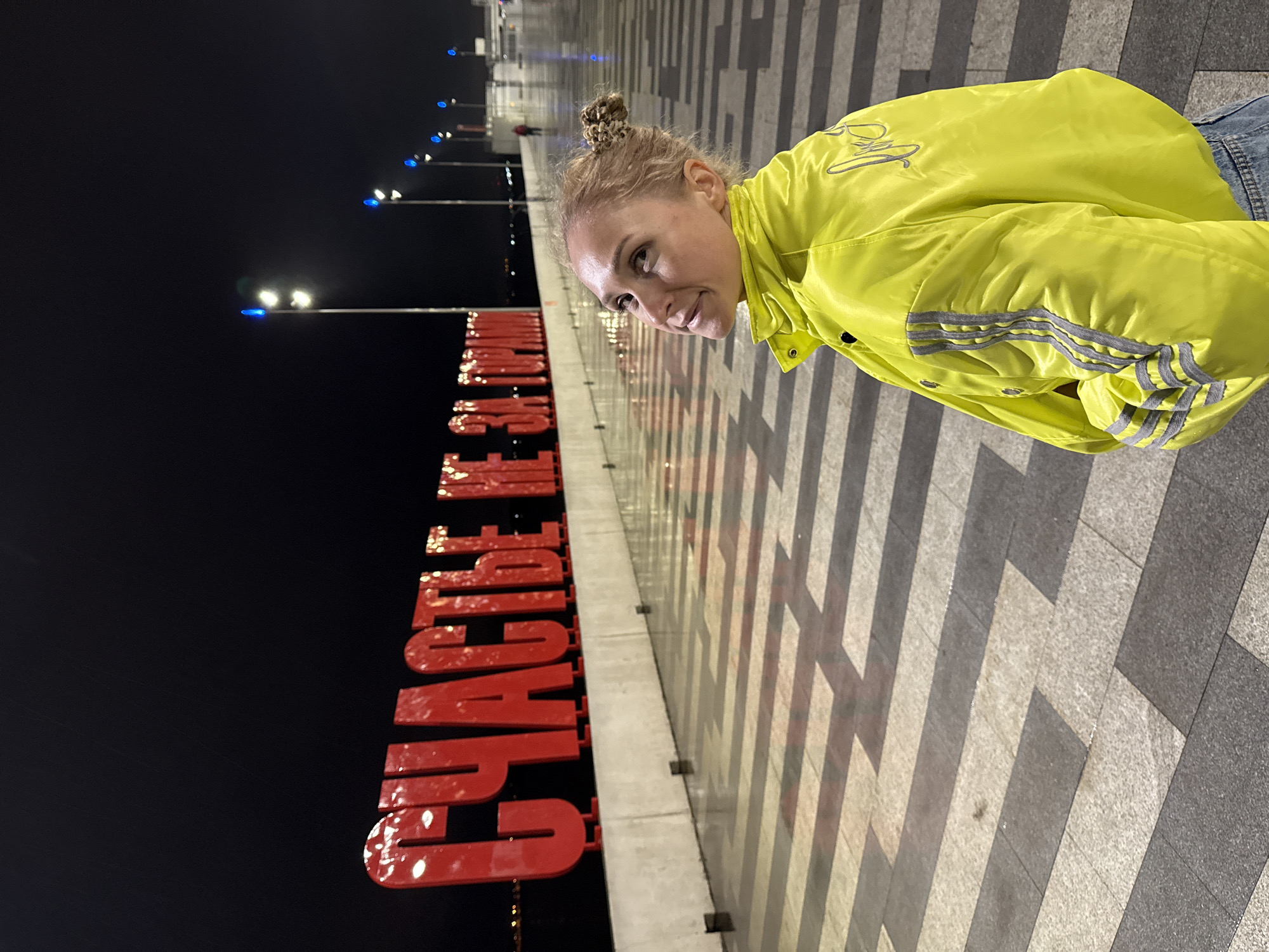 A vibrant nighttime outdoor photograph captures a woman posing next to a prominent red art installation in Perm, Russia. The woman, with light hair styled in a bun, is dressed in a bright neon yellow jacket featuring three grey stripes on the sleeves and blue jeans. She looks towards the viewer with a gentle smile, her head slightly tilted, conveying a cheerful demeanor.
Behind her, to the left, stands a large, three-dimensional red sign with glossy letters, spelling out "СЧАСТЬЕ НЕ ЗА ГОРАМИ." This Russian phrase translates to "Happiness is not far off" and is a well-known art object in Perm. The sign's illuminated surface and the distant streetlights cast reflections on the wet-looking paved ground. The ground is composed of geometric patterns of light and dark grey rectangular tiles.
The background is a dark night sky, punctuated by rows of distant streetlights emitting blue and white glows. The reflections on the ground suggest a damp surface, possibly from recent rain or dew. The scene depicts a lively embankment or promenade at night, with the iconic sign serving as a distinct landmark in Perm.
