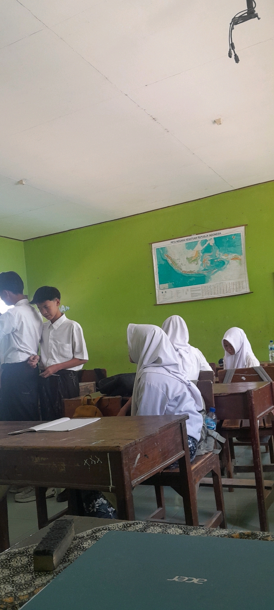 This image depicts a classroom scene, likely in Tahunan, Indonesia, with several students present. Two male students in white shirts and dark pants are standing on the left side of the frame, facing each other in conversation. One of them appears to be looking down at the other. To the right, three female students, all wearing white hijabs and uniforms, are seated at desks. One is looking towards the left, another is facing away from the camera, and the third is looking down. The room has light green walls with a map of Indonesia hanging on the right wall. Desks and chairs are made of wood. In the foreground, a wooden desk has an open notebook and what appears to be an eraser or chalk holder. A laptop with the "acer" logo is visible on another desk in the lower foreground. The ceiling is white with visible seams and some minor discoloration. There are electrical conduits visible on the ceiling in the upper right.