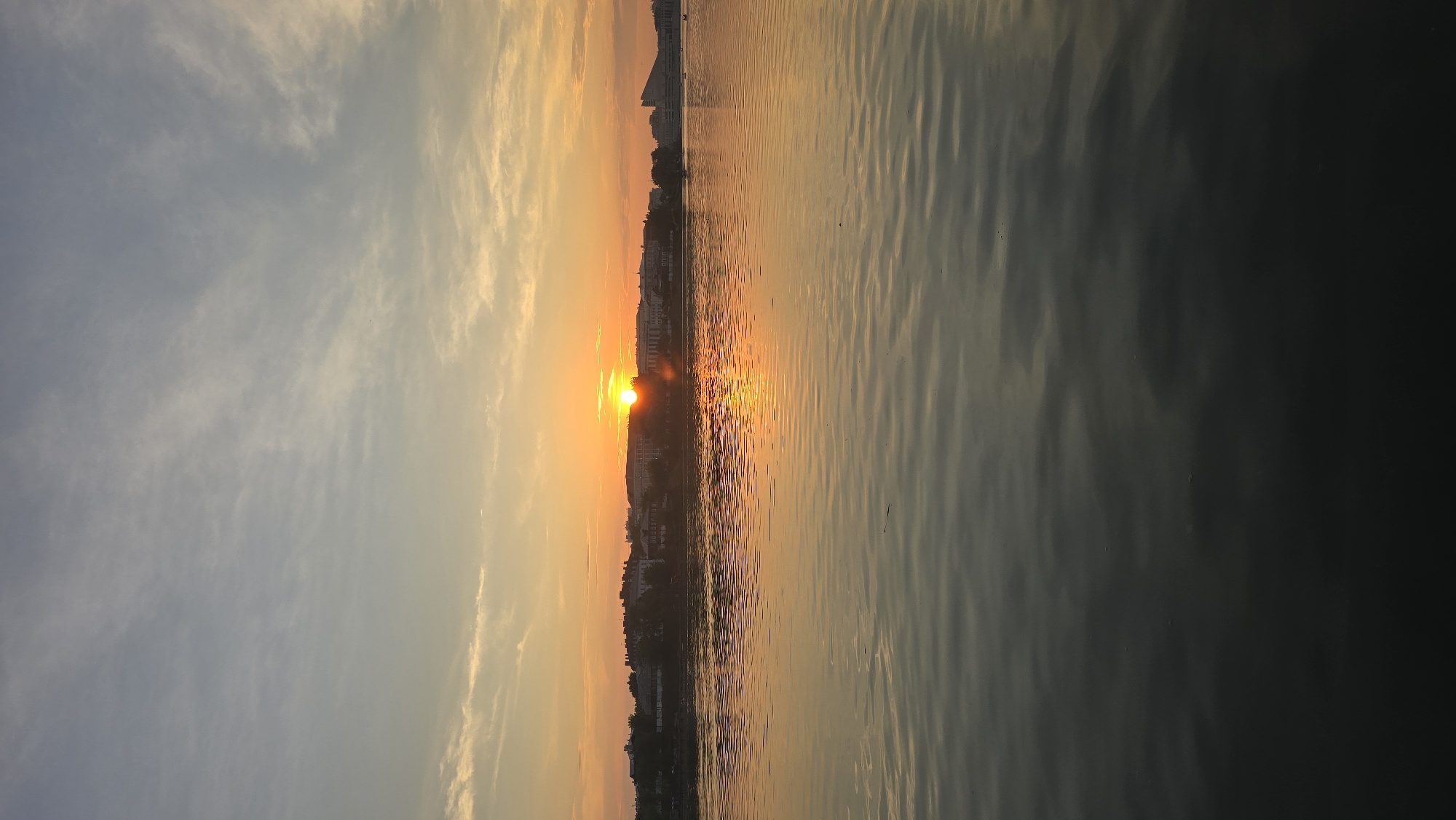 A vertical, medium shot captures a serene sunset over the Kazanka River in Kazan, Russia. The sun, a bright orb, is partially obscured by a silhouetted line of buildings and trees on the distant bank, casting a warm orange glow across the sky and reflecting shimmering patterns on the water's surface. The sky is a gradient of pale blue and soft orange, with wispy clouds scattered throughout. The river occupies the lower half of the frame, its dark, rippling surface reflecting the colors of the sunset. No people or specific actions are discernible in this tranquil scene.