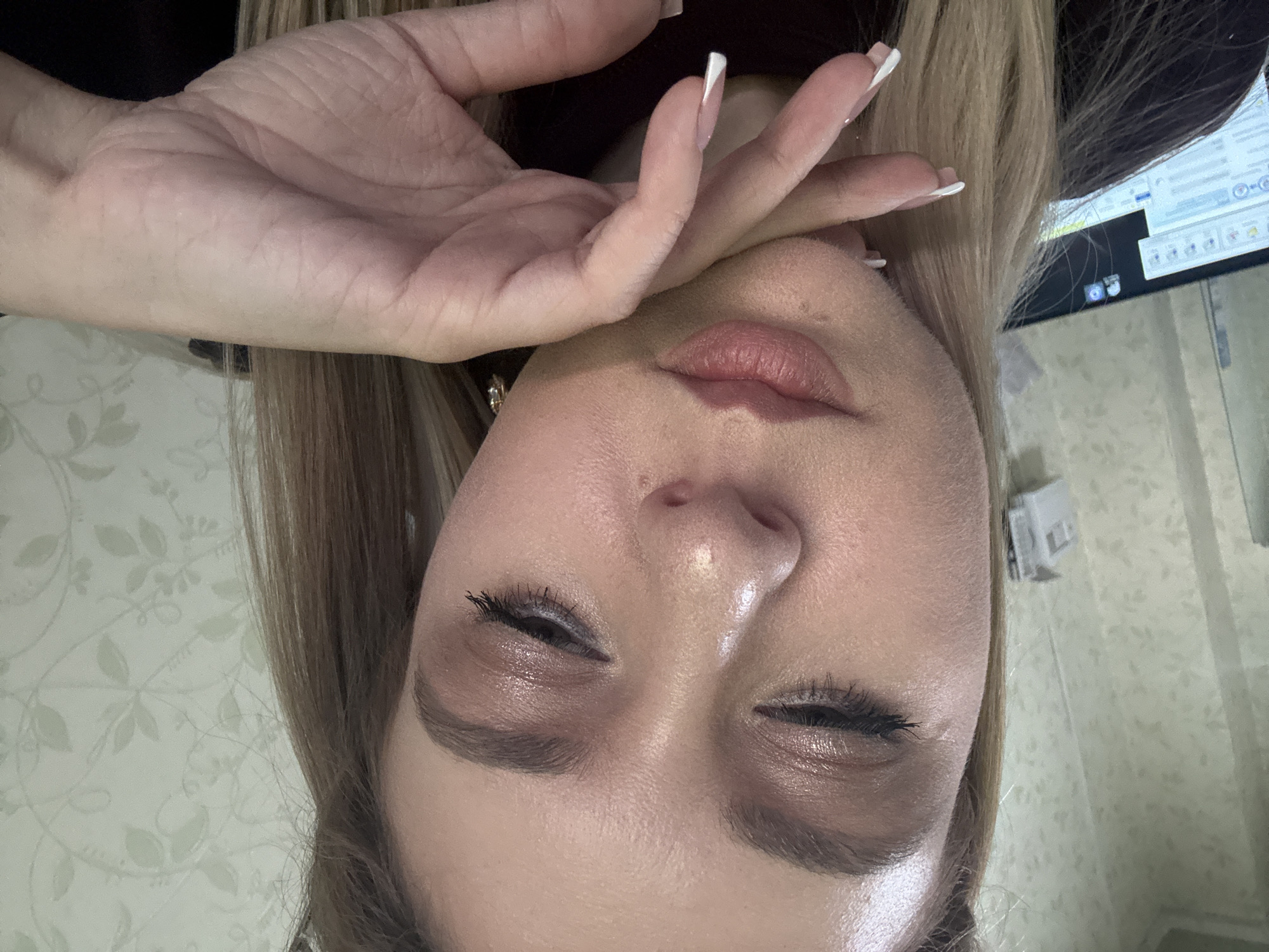 A woman with blonde hair and makeup is captured in a close-up shot, her face angled downwards with her eyes closed. Her right hand is raised to her chin, with her fingernails painted white. She is wearing a dark shirt. The background features a wall with a floral pattern and a computer monitor visible on the left side of the frame. The lighting appears to be artificial and directed towards her face. The scene is indoors and appears to be a personal or home setting.