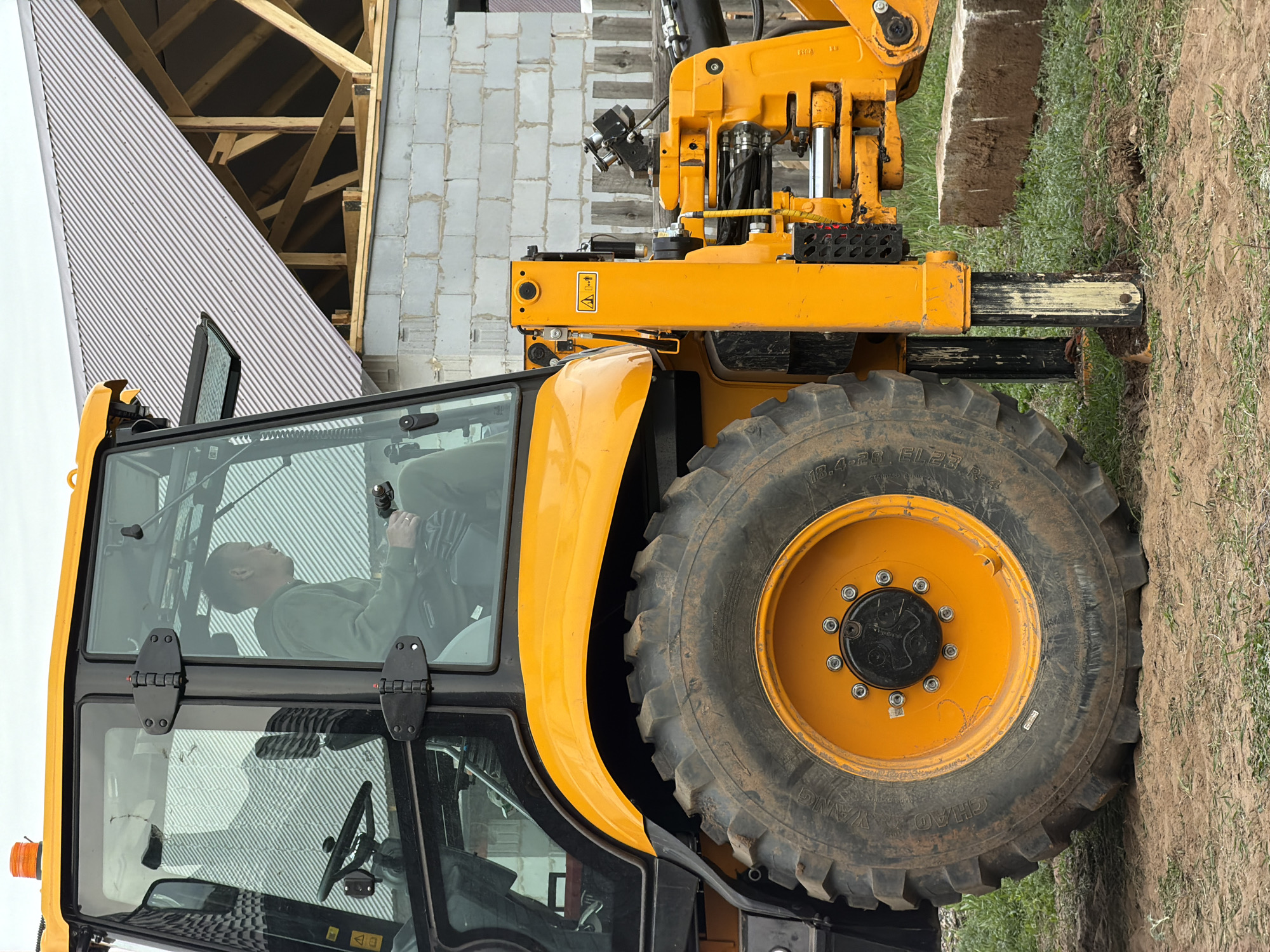 The image shows a yellow JCB telehandler parked on a dirt and grass surface. The telehandler is positioned next to a building under construction, evidenced by exposed wooden framing and gray brick walls. Inside the cab, a man is visible, seated in the driver's seat. The scene appears to be in a rural or semi-rural environment. Location information (Chaikovsky, Russia) is provided externally.